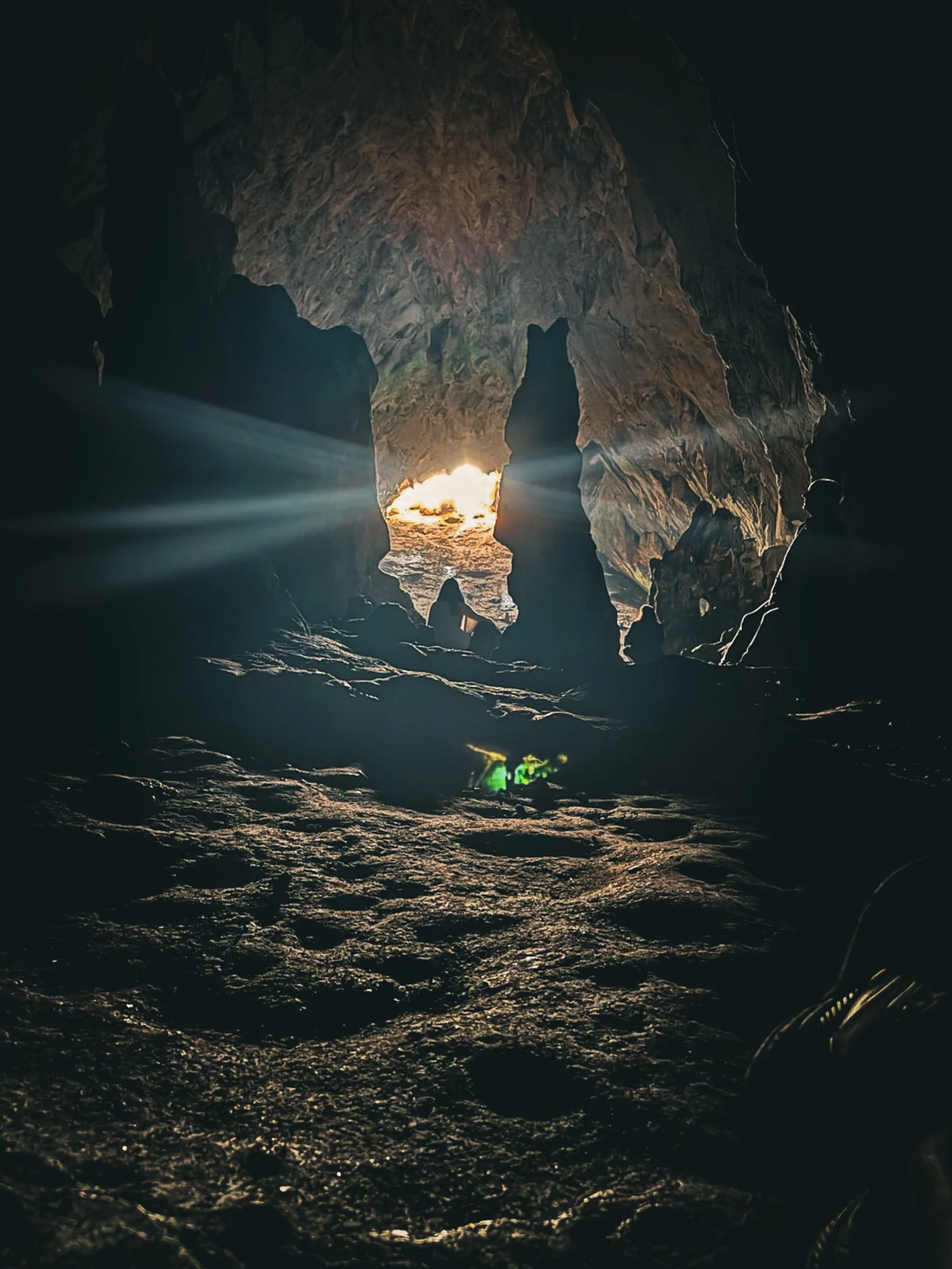 Light beams pierce through a narrow cave opening into deep darkness, illuminating the cave floor in shafts of gold