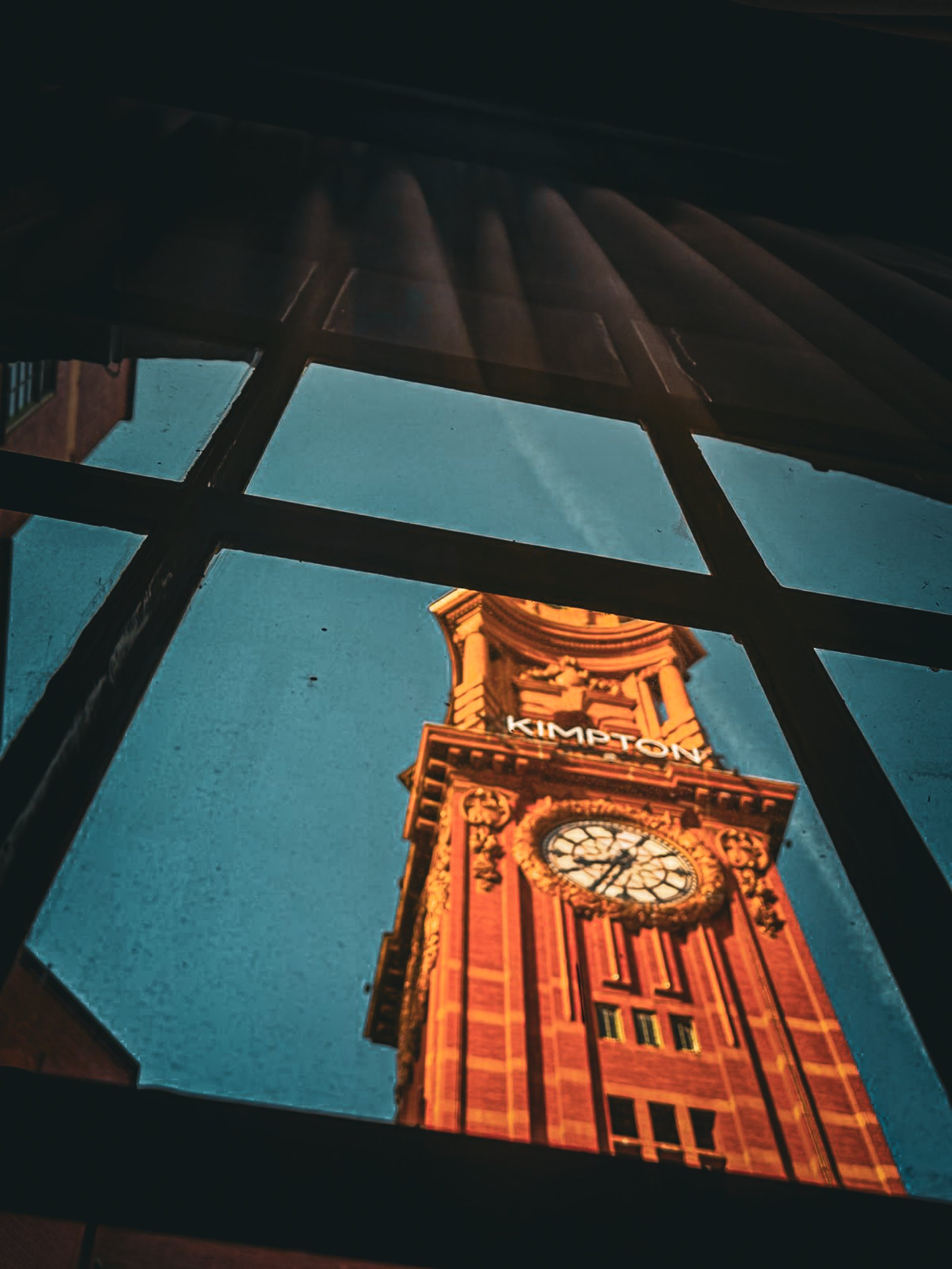 A Victorian clock tower bearing the Kimpton sign, photographed upward through a grid of glass panes, glowing amber against a blue sky