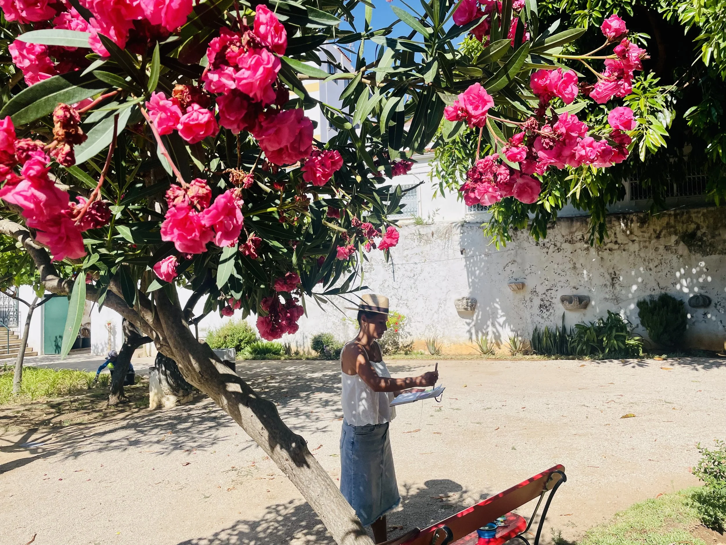 A woman in a white top sketching in her sketch book under the bright pink flowers in the sunny courtyard.