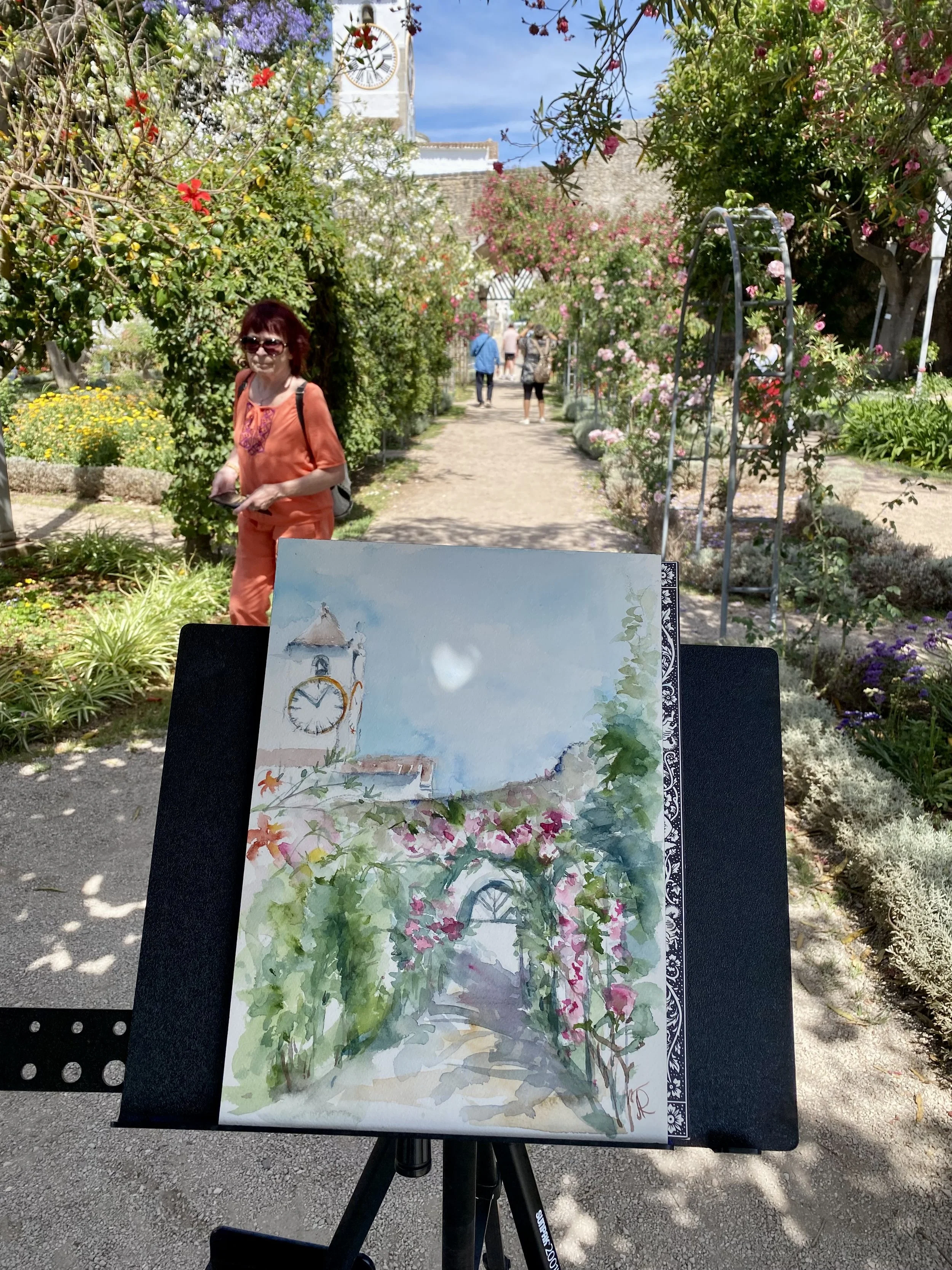 A watercolor painting on an easel depicts a clock tower with a light blue sky background, surrounded by pink and purple flowers in a garden. The scene is outdoors, with trees and a path leading toward the tower.