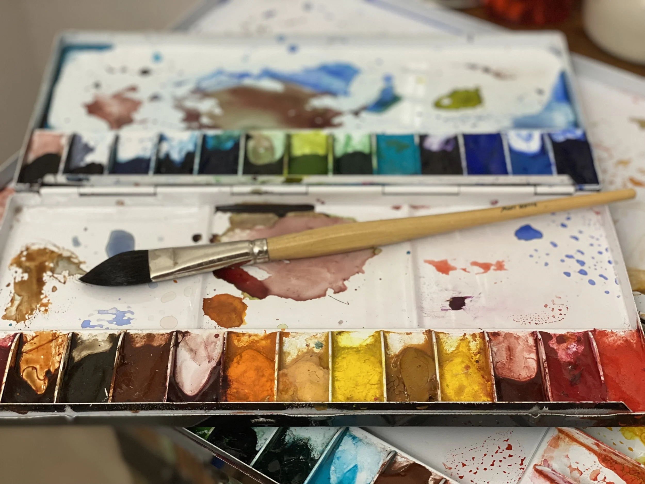 A watercolor paint set with various colors, a paintbrush resting on the palette, and some paint splatters around.