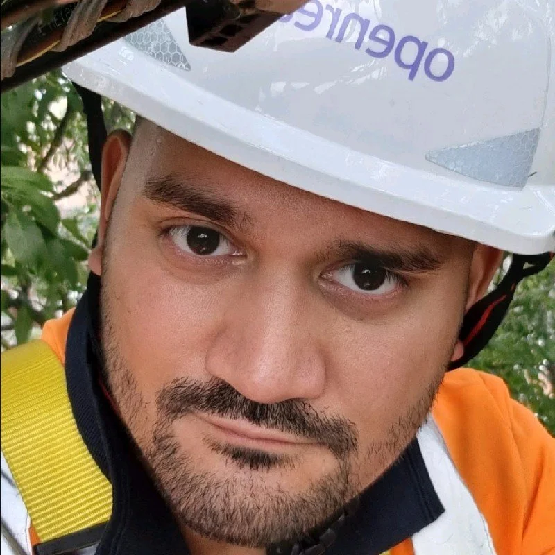 Close-up of a man wearing a white safety helmet with the word 'operate' visible, and an orange safety vest, outdoors with green foliage in the background.