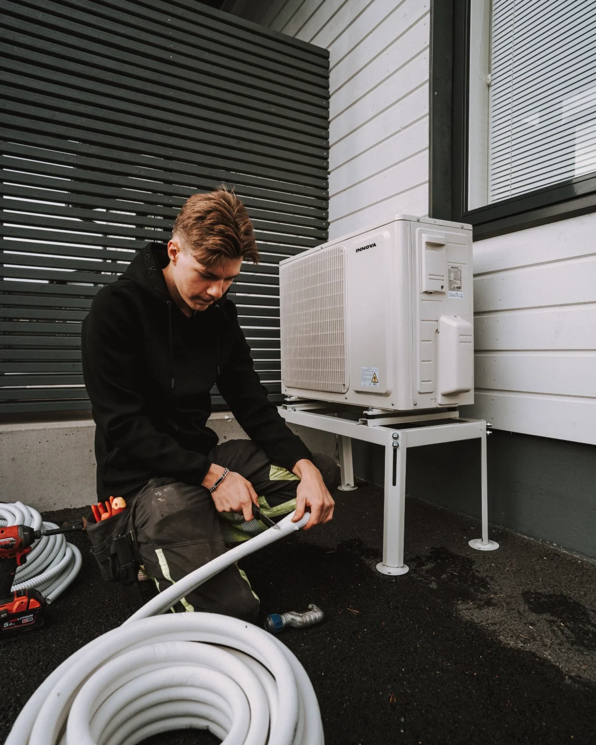 Nuori mies asentaa tai huoltaa ilmalämpöpumppua ulkona rakennuksen vieressä, lattialla on letku ja työkaluja.