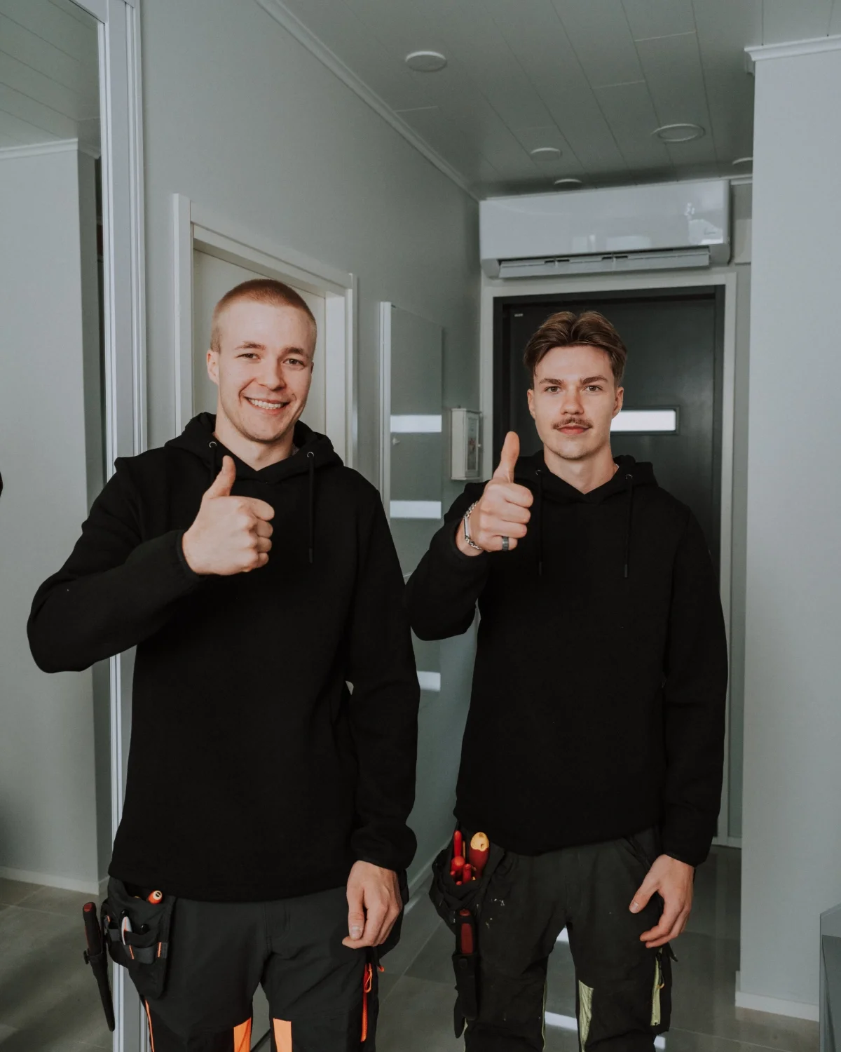 Kaksi nuorta miestä pukeutuneena mustiin huppareihin ja työvaatteisiin, hymyilevät ja antavat peukkuja katsojalle sisätiloissa.