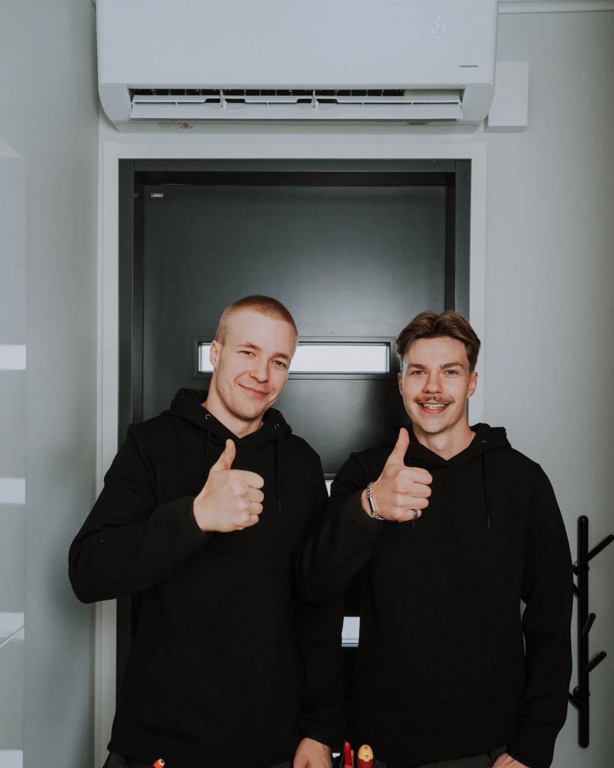 Kaksi miestä hymyilevät ja vilkuttelevat valkoisella taustalla, heidän päälleen on pukevat mustat hupparit.