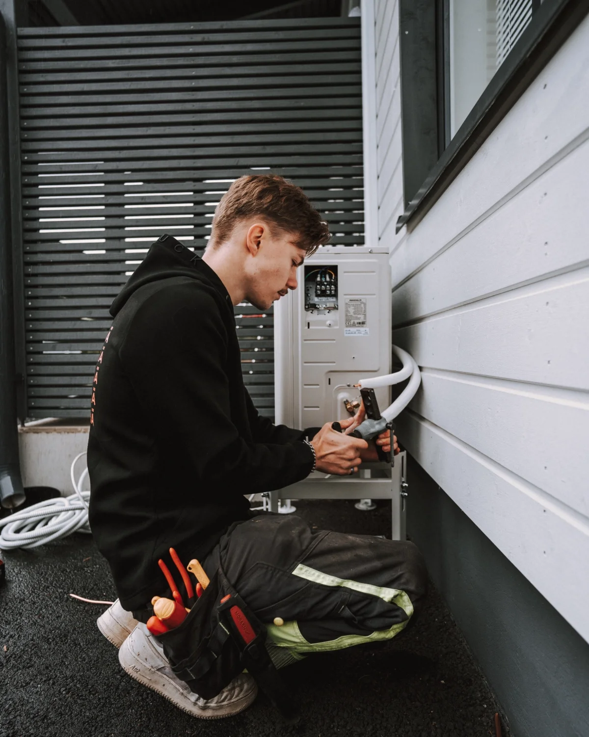 Nuori mies työskentelee sähköasennuksissa ulkona, istahtaa ja käyttää työkaluja ilmastointilaitteen tai sähkölaitteen huoltoon.