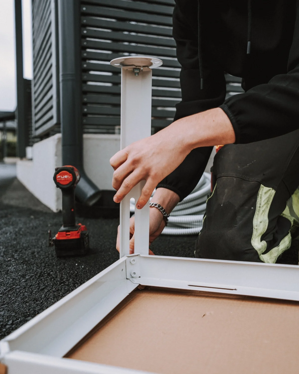 Henkilö kiinnittää kalustetta ulkona, taustalla näkyy työkalupistooli ja rakennuksen ilmanvaihtoventtiileitä.