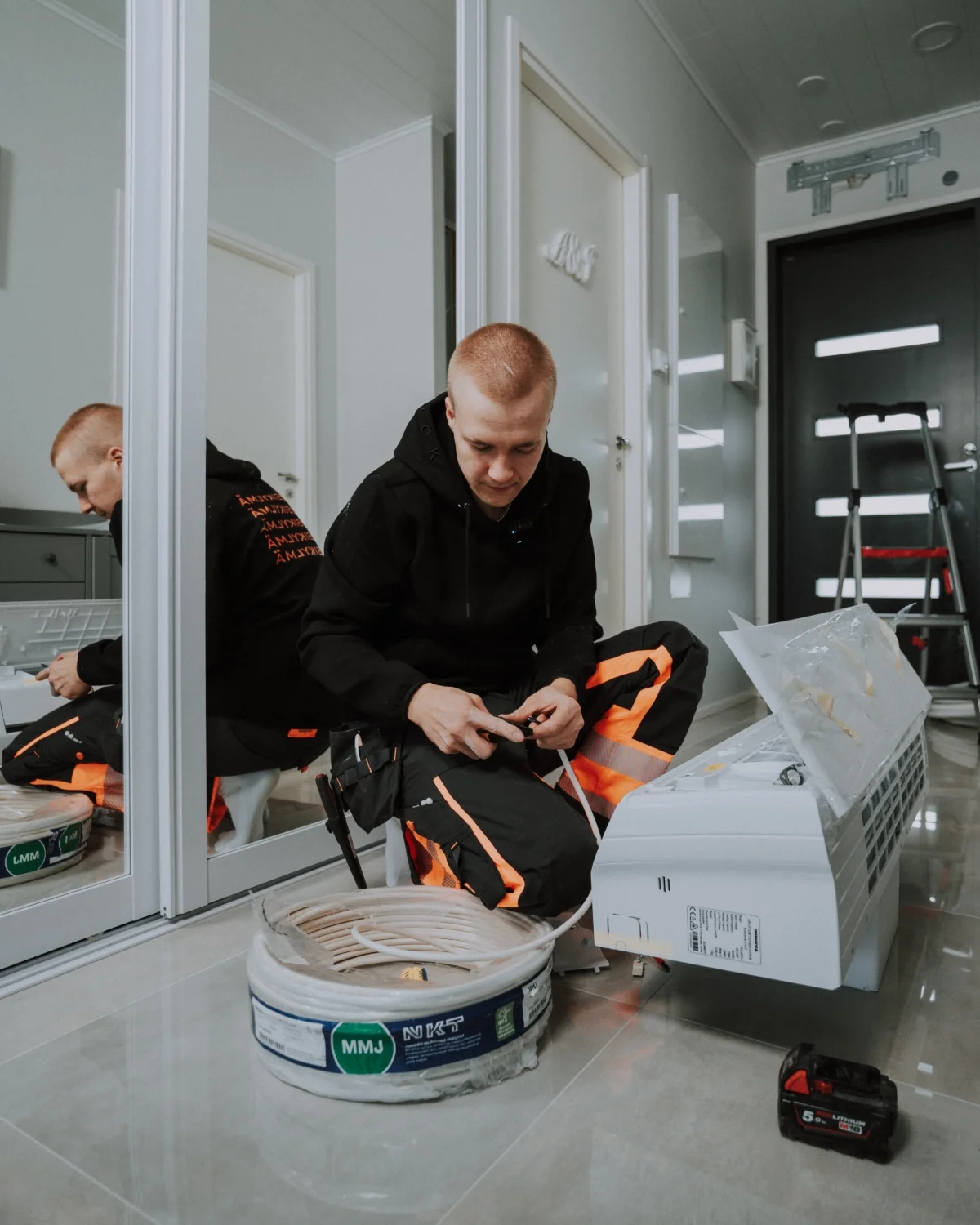 Mies työskentelee sähköasennustyön parissa sisällä, istuen lattialla ja kytkien johtoja, lähellä ilmastointilaitetta ja työkalupakkia.