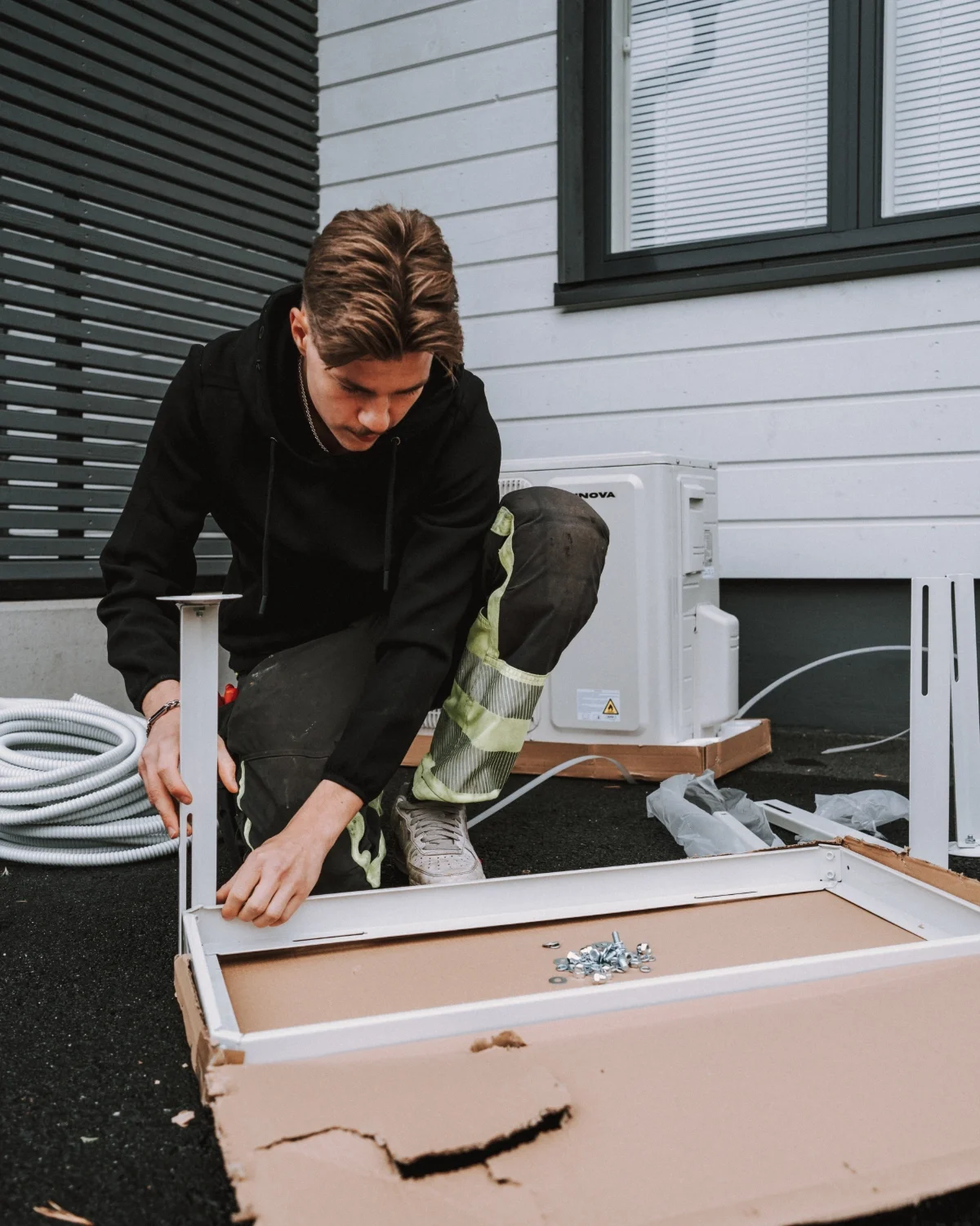 Nuori mies rakentaa tai asentaa valkoinen kaluste, mahdollisesti pöytää tai tasoa, ulkoalueella. Hän on kumisaappaissa ja roskakuvussa näkyy metalliruuveja. Taustalla valkoinen ulkoseinä ja ikkunat.