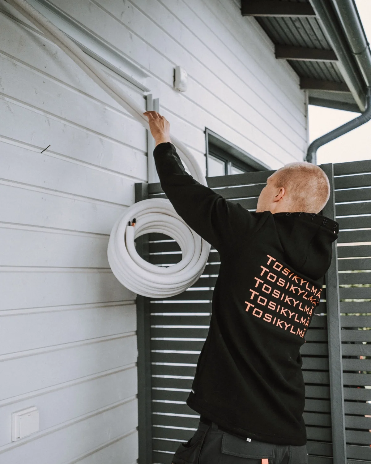 Mies asennamassa ulkoilmarenkaita seinälle, istuvalta tai seisovasta asennosta, ulko-alueella, takaseinän vieressä grillin tai patio asiakkaan. Hänellä on musta huppari, jossa on teksti 'TOSIKYLMÄ' toistuvasti selässä, ja hän ripustaa valkoisia letku