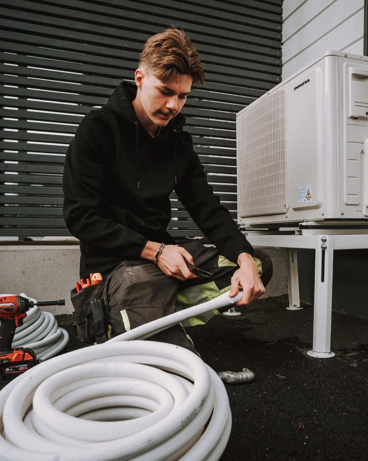 Mies henkilö työskentelee ulkona, kääriä jännitettä joustavaan letkuun, ympärillä on ilmastointilaitteen ulkoyksikkö ja työkalut.