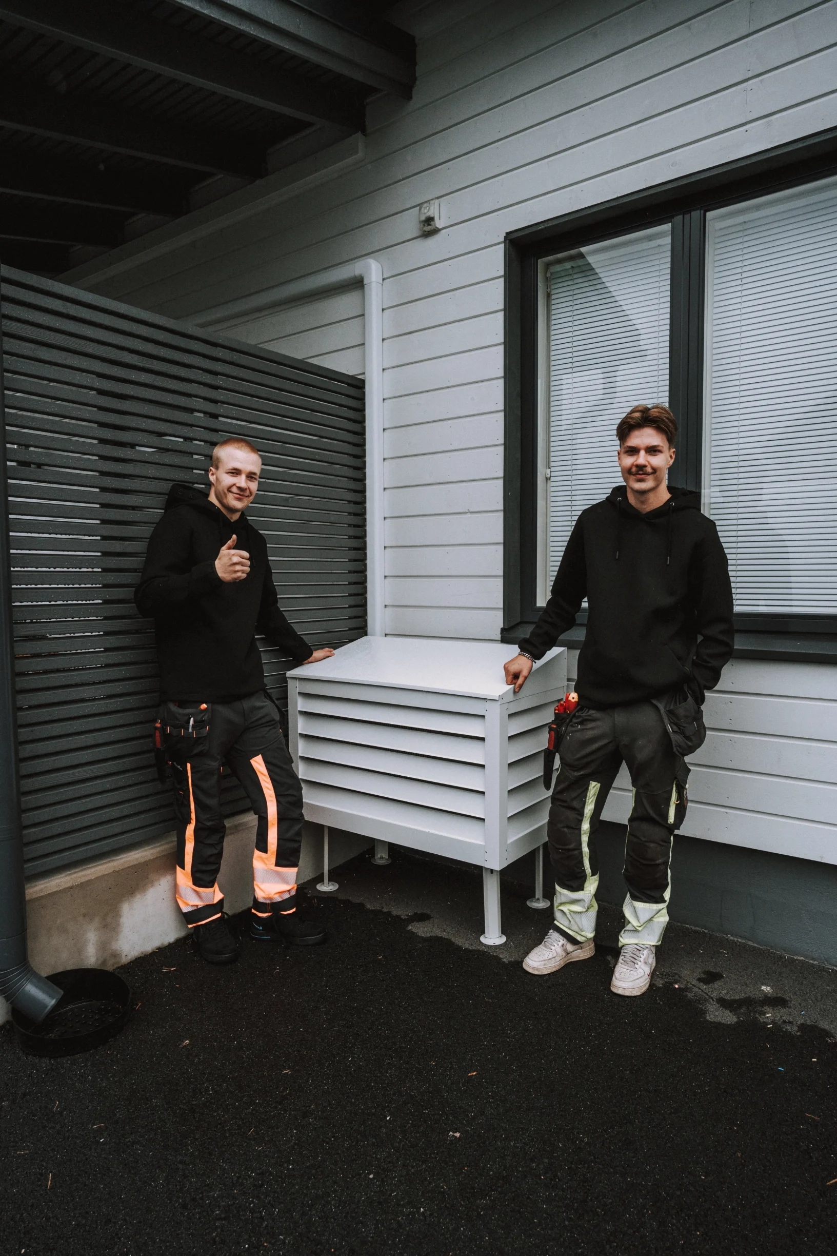 Kaksi miestä seisoo pihalla rakennuksen seinämän vieressä, toinen antaa peukkua näytteenä, molemmilla työkalut vyöissään.