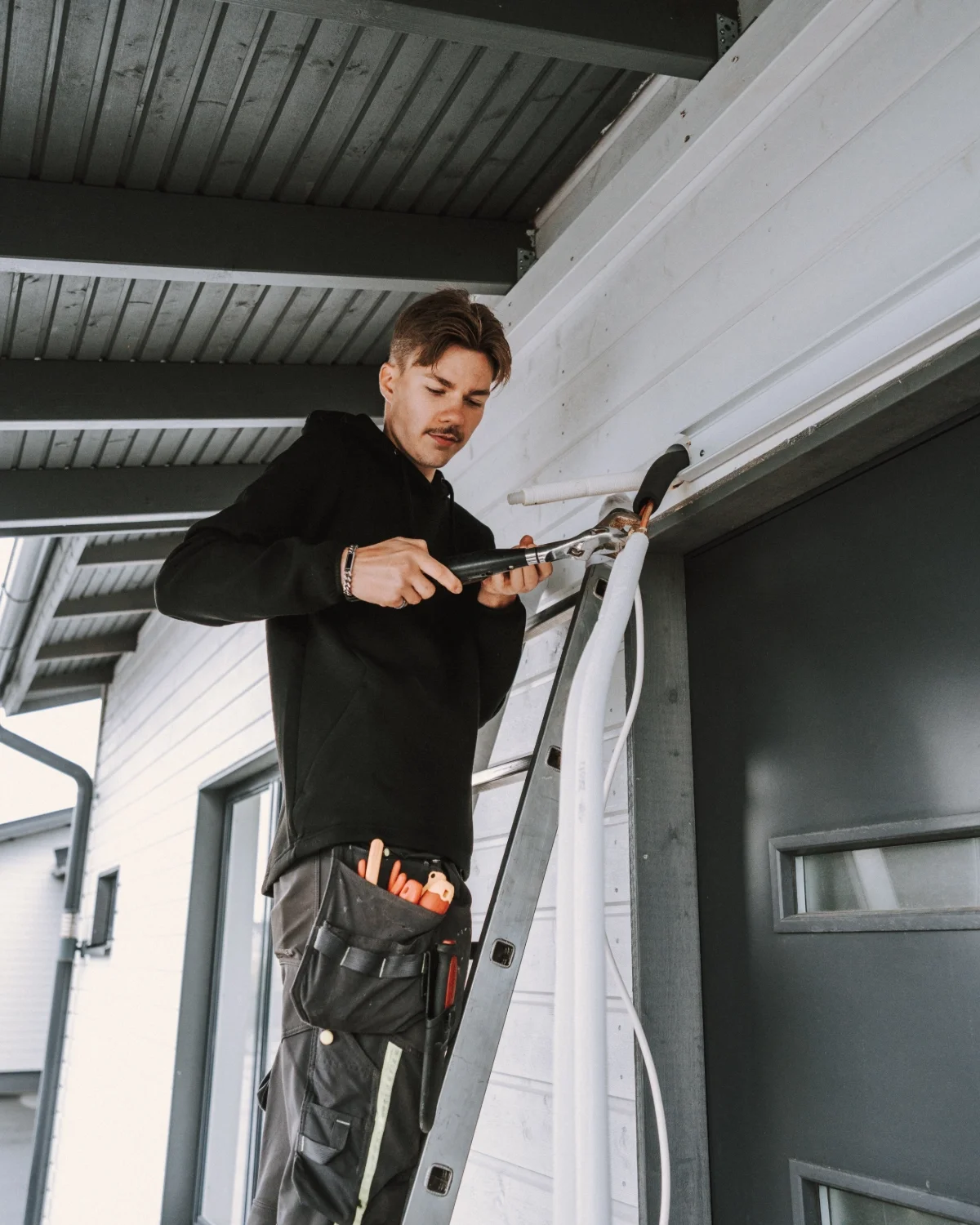 Mies tekee sähköasennustöitä, hän seisoo tikkailla ja käyttää työkaluja talon ulkoseinän sähköjohtojen asentamiseen.