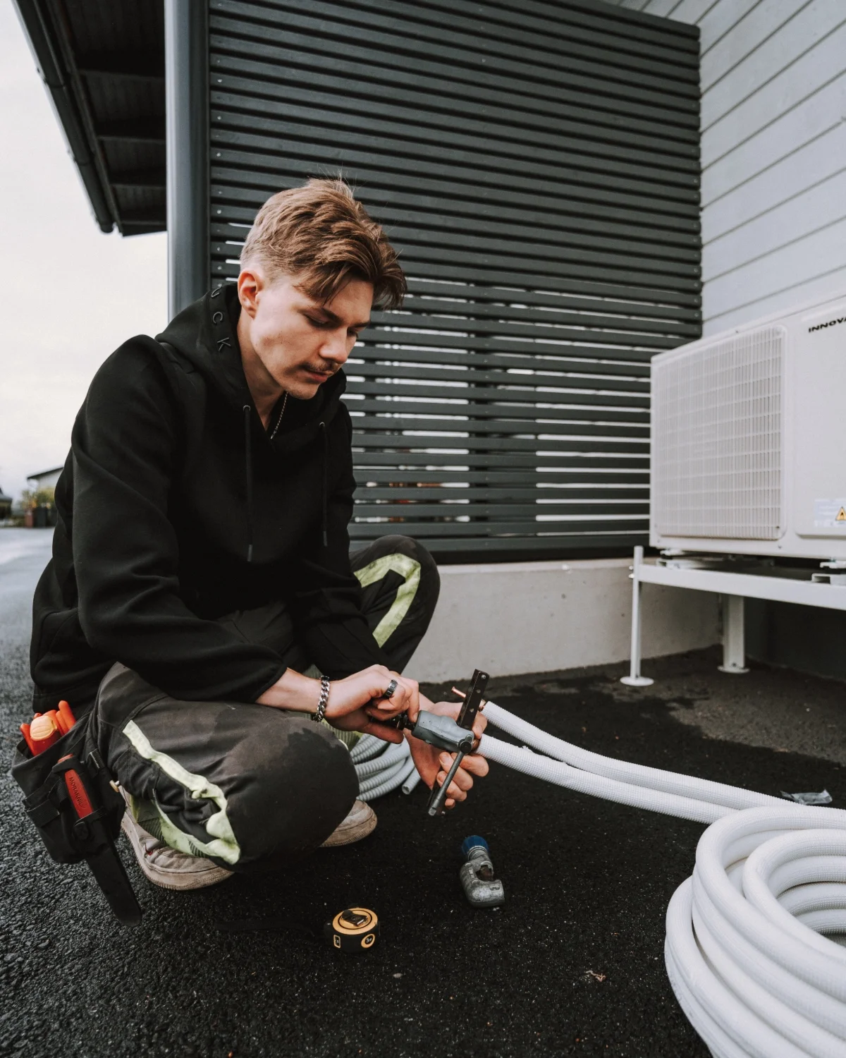 Nuori mies työskentelee ilmastointilaitteen huollossa ulkona, käyttää mittaustyökalua ja mittanauhaa.