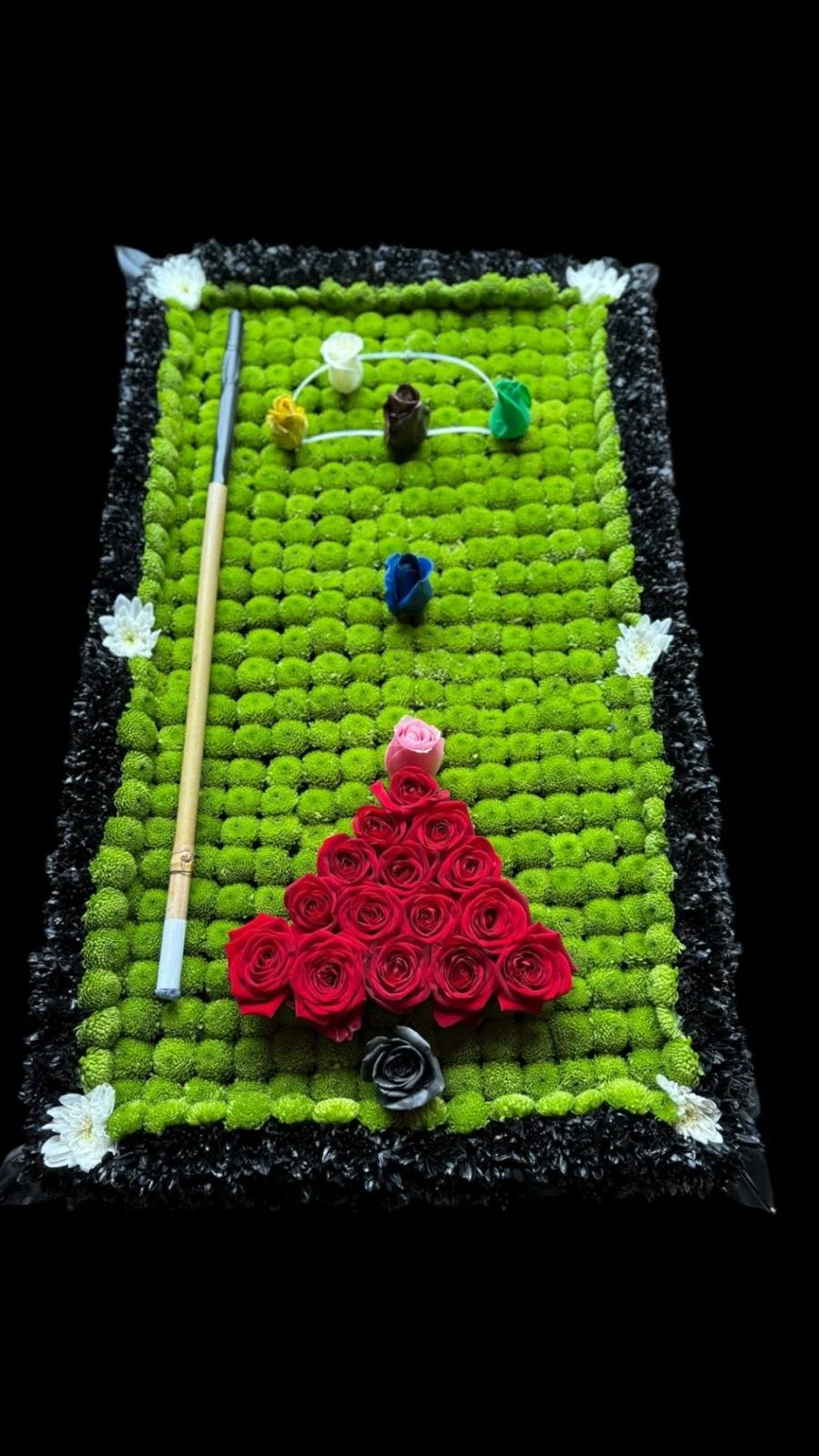 A decorated cake resembling a soccer field with green grass-like frosting, a black border, white flowers at the corners, and a triangle of pink and red roses at the bottom. It has a miniature soccer goal made of white string, some small colorful ball