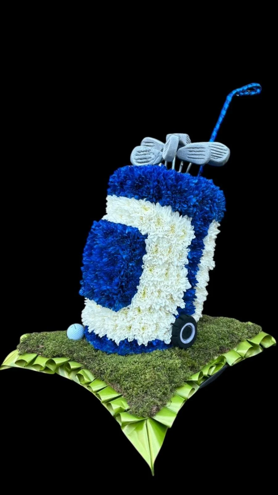 A floral sculpture of a golf bag with clubs, made from various colored flowers, placed on a green leaf base against a black background.