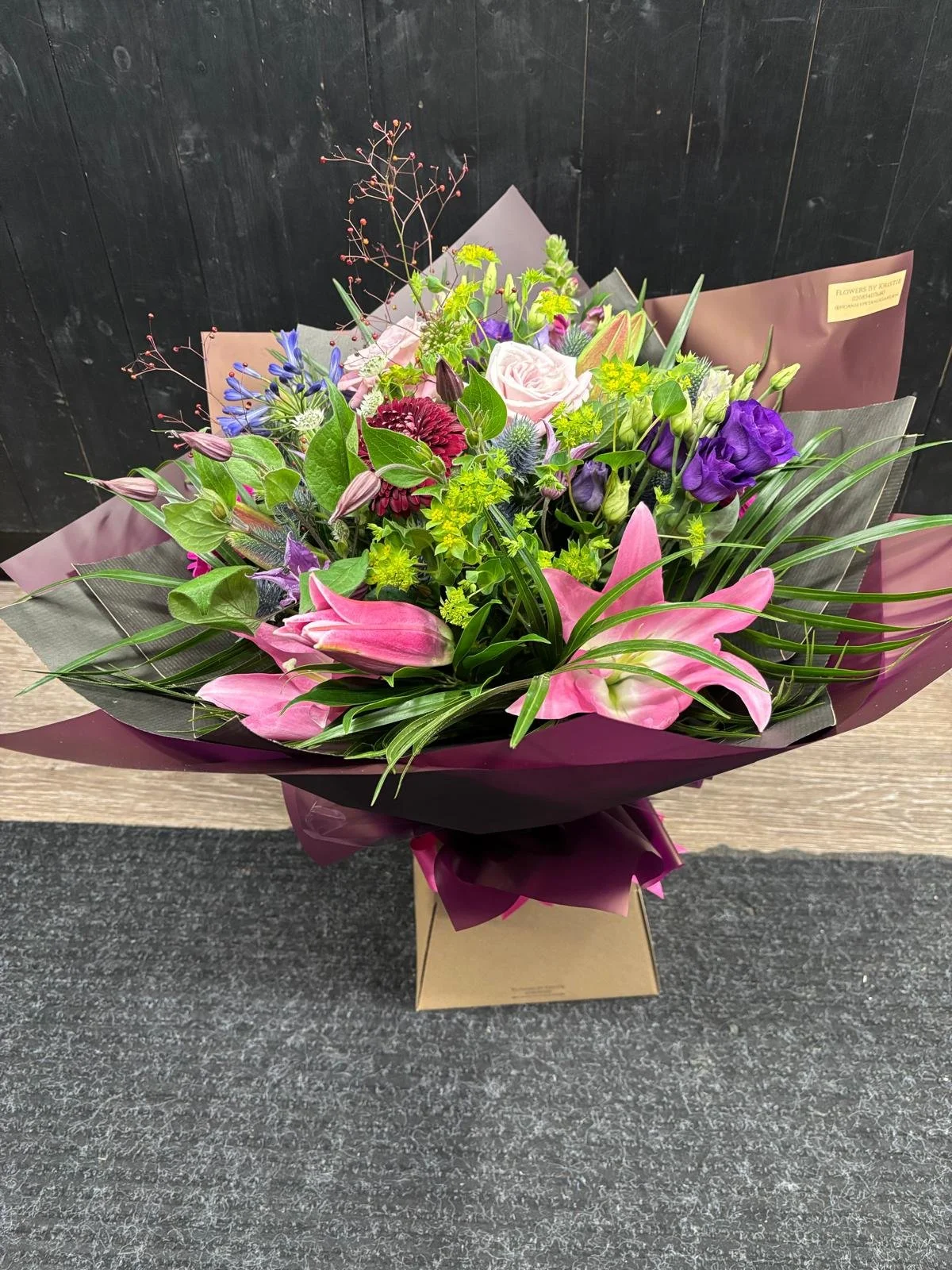 A colorful bouquet of pink lilies, purple flowers, and various greenery wrapped in purple and gray paper, sitting on a beige box on a dark surface with a black wall in the background.