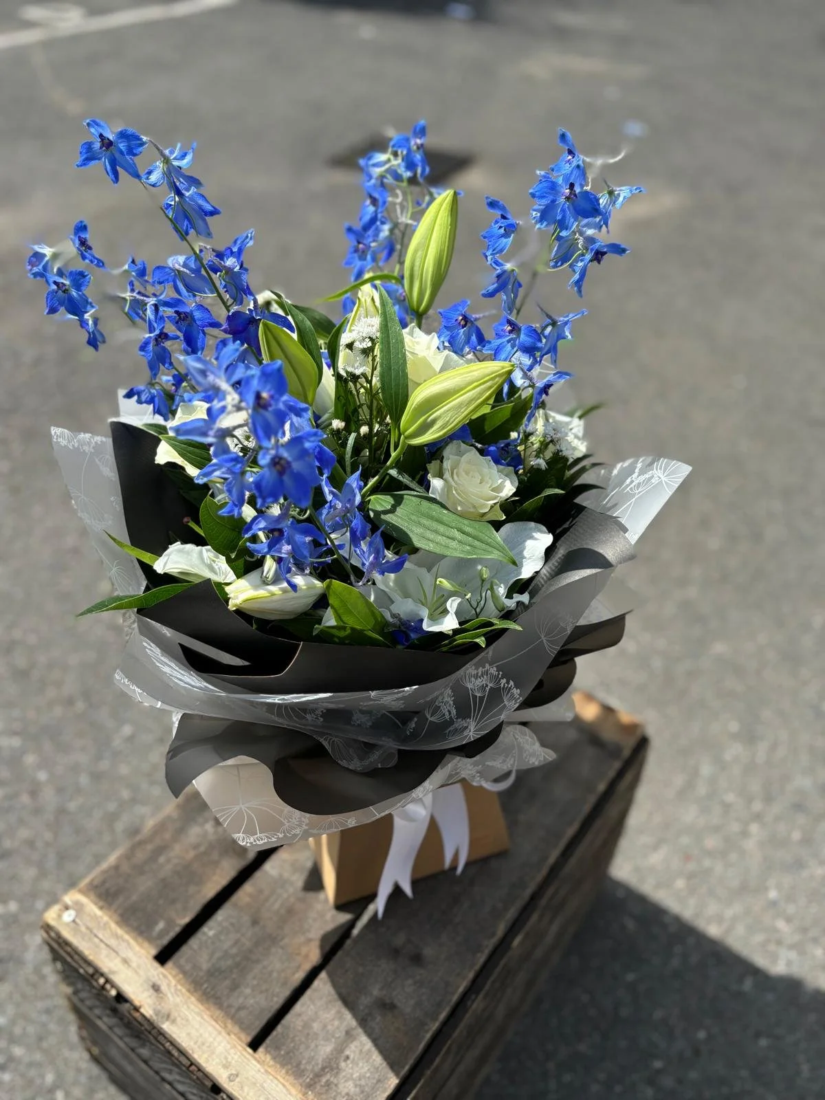 A bouquet of blue, white, and green flowers wrapped in black and white paper, placed on a wooden crate outdoors.