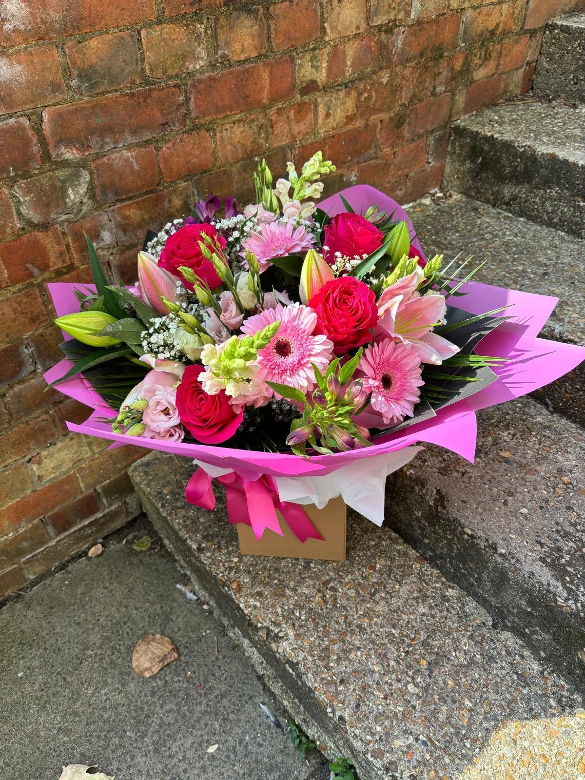 A bouquet of pink, red, and white flowers with green leaves, wrapped in pink and white paper, sitting on a concrete step next to a brick wall.