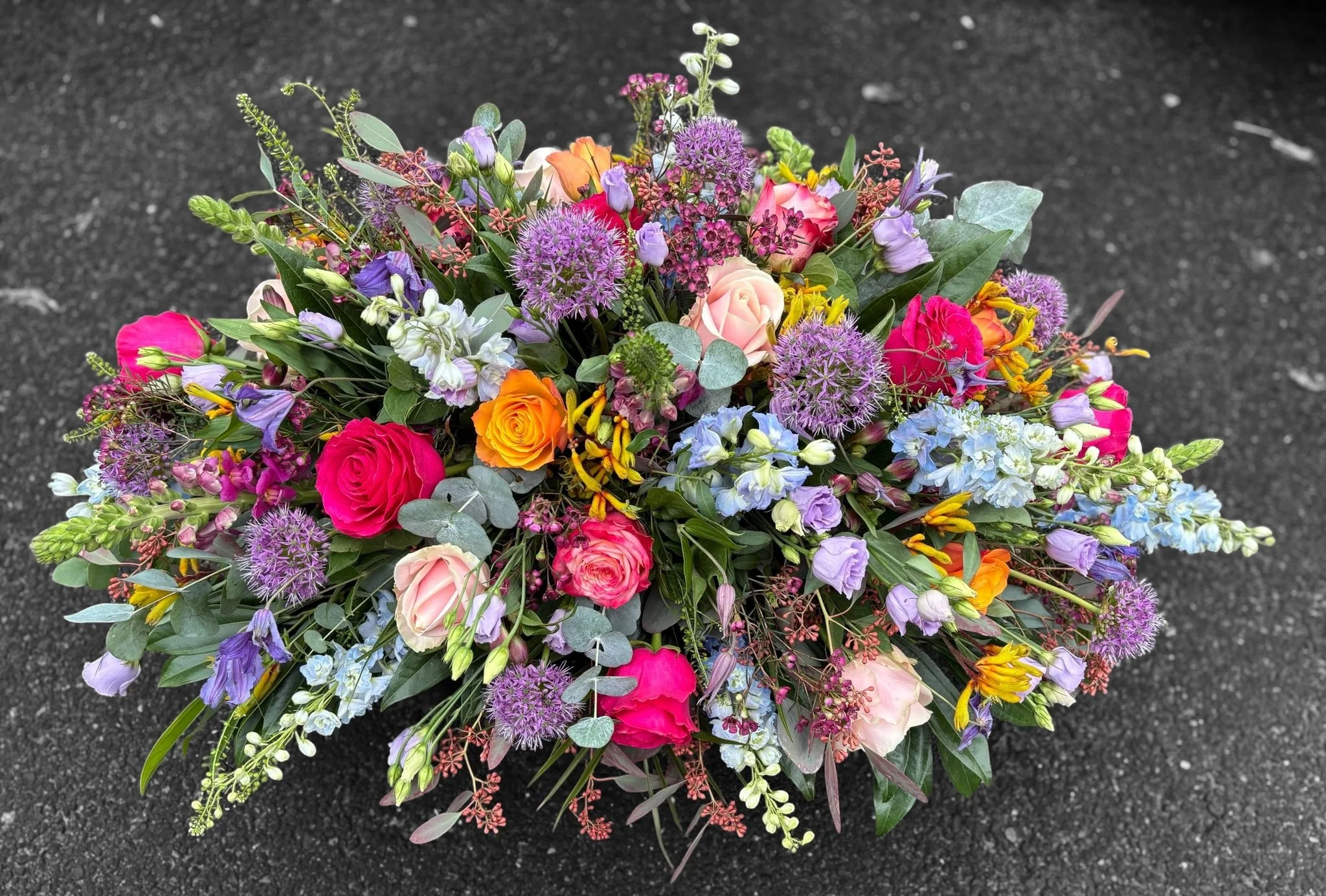 A colorful bouquet of various flowers including roses, purple alliums, and yellow blossoms, arranged on a dark surface.