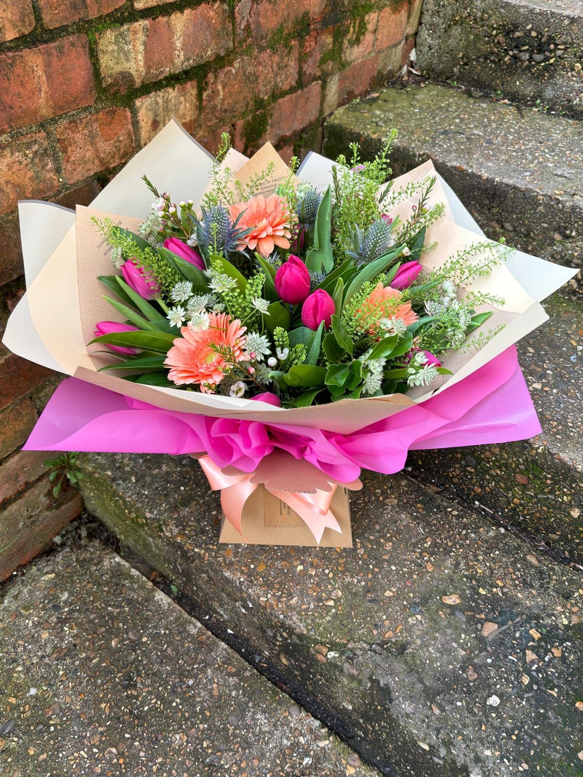 A colorful bouquet of pink, peach, and white flowers wrapped in pink and beige paper with a pink ribbon sits on a mossy brick step.