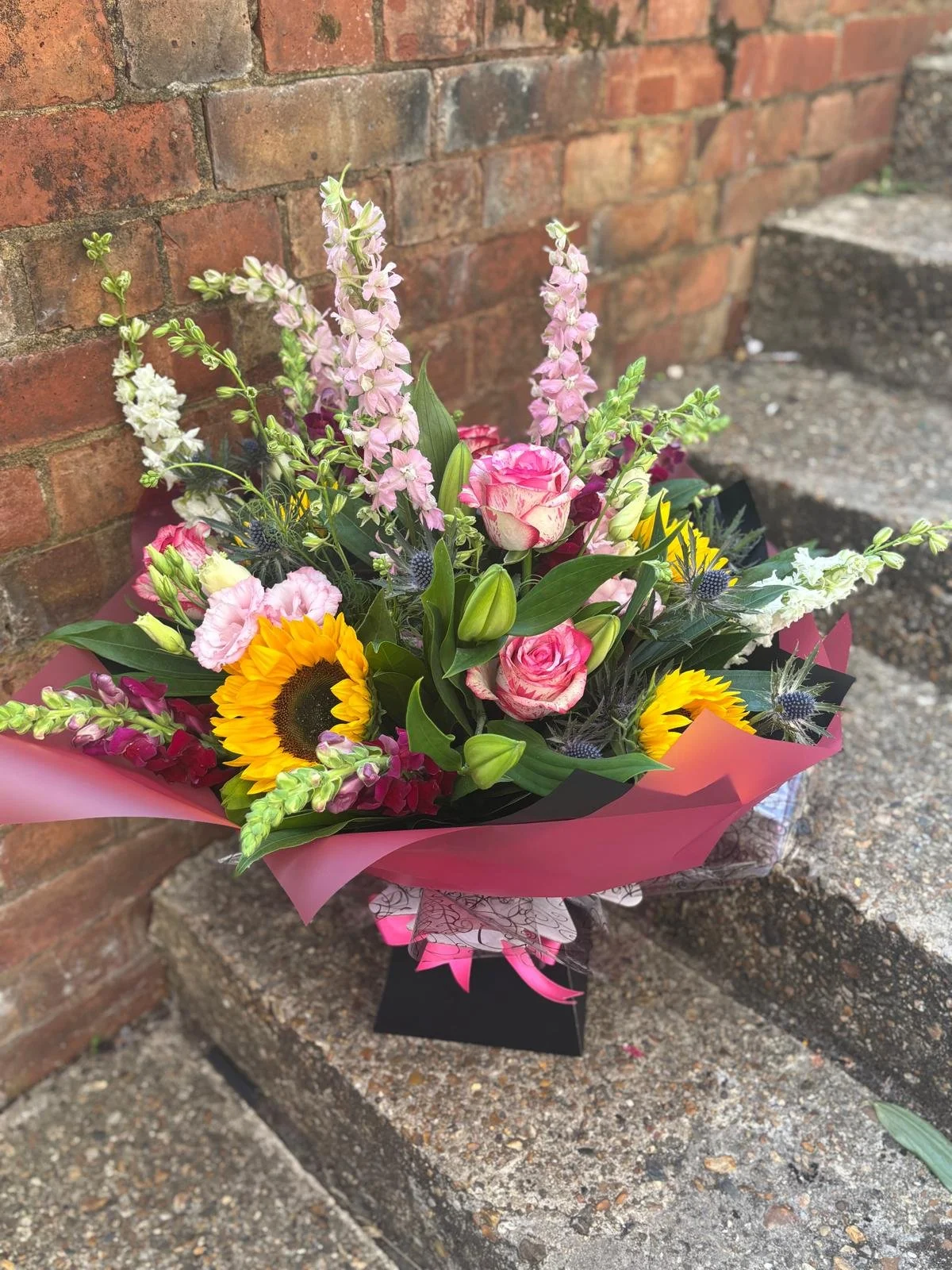 A colorful bouquet of mixed flowers including pink roses, sunflowers, white snaps, and purple thistles, wrapped in pink paper and placed on concrete steps against a brick wall.