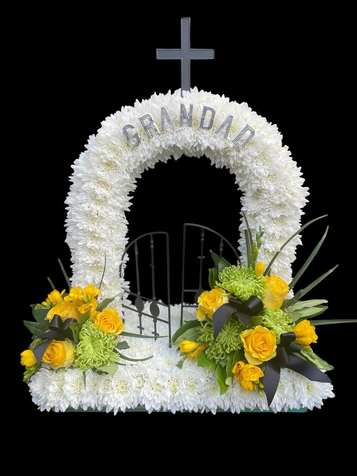 Funeral floral display with white flowers, yellow roses, green flowers, and a black ribbon, with a cross on top and a sign that says "Grandad."
