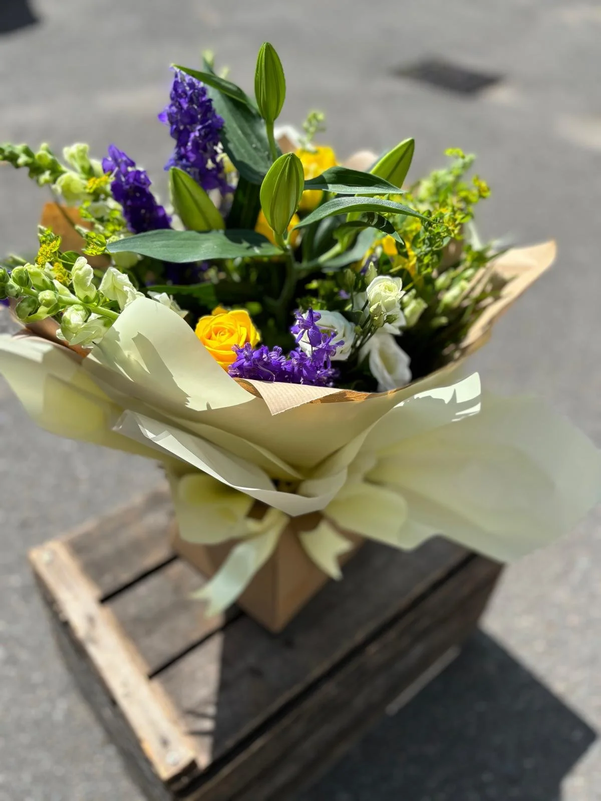 A bouquet of colorful flowers wrapped in paper, placed in a wooden crate on a paved surface.