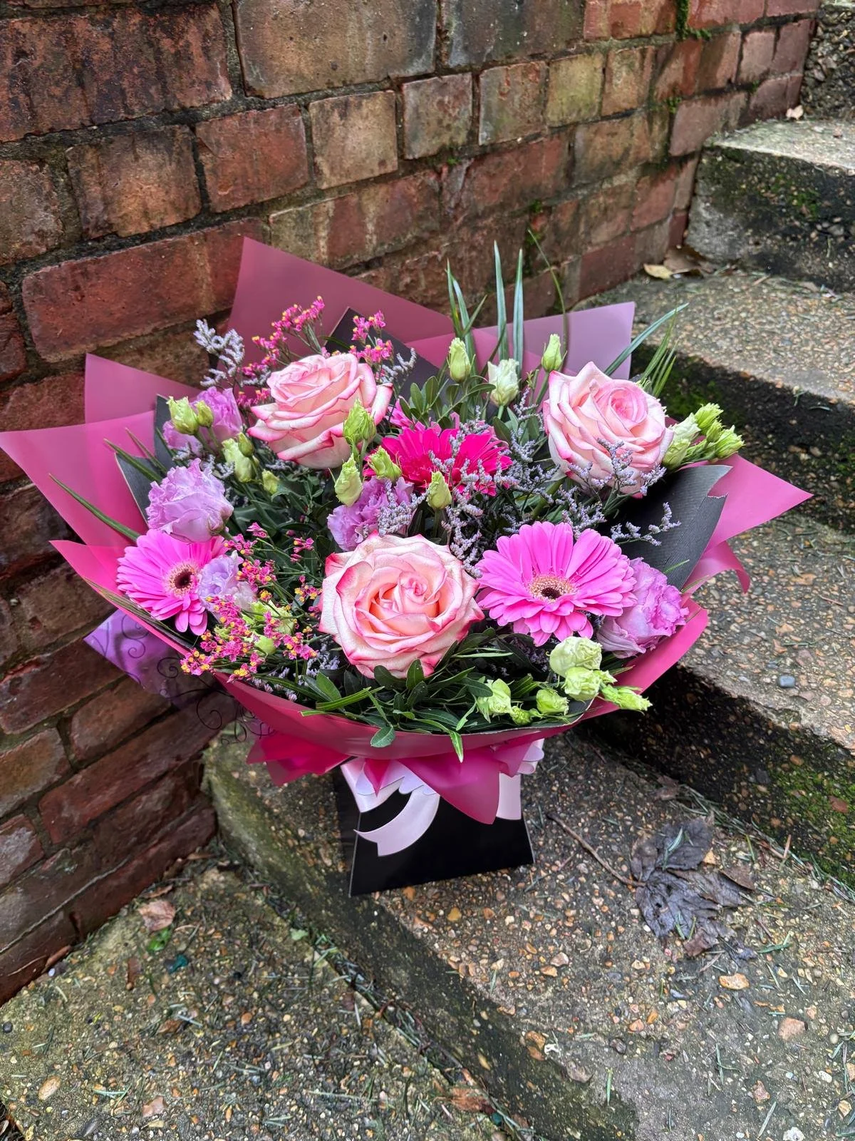 A bouquet of pink roses, pink gerbera daisies, light purple lisianthus, and assorted greenery wrapped in pink and purple paper, placed on a concrete step against a brick wall.