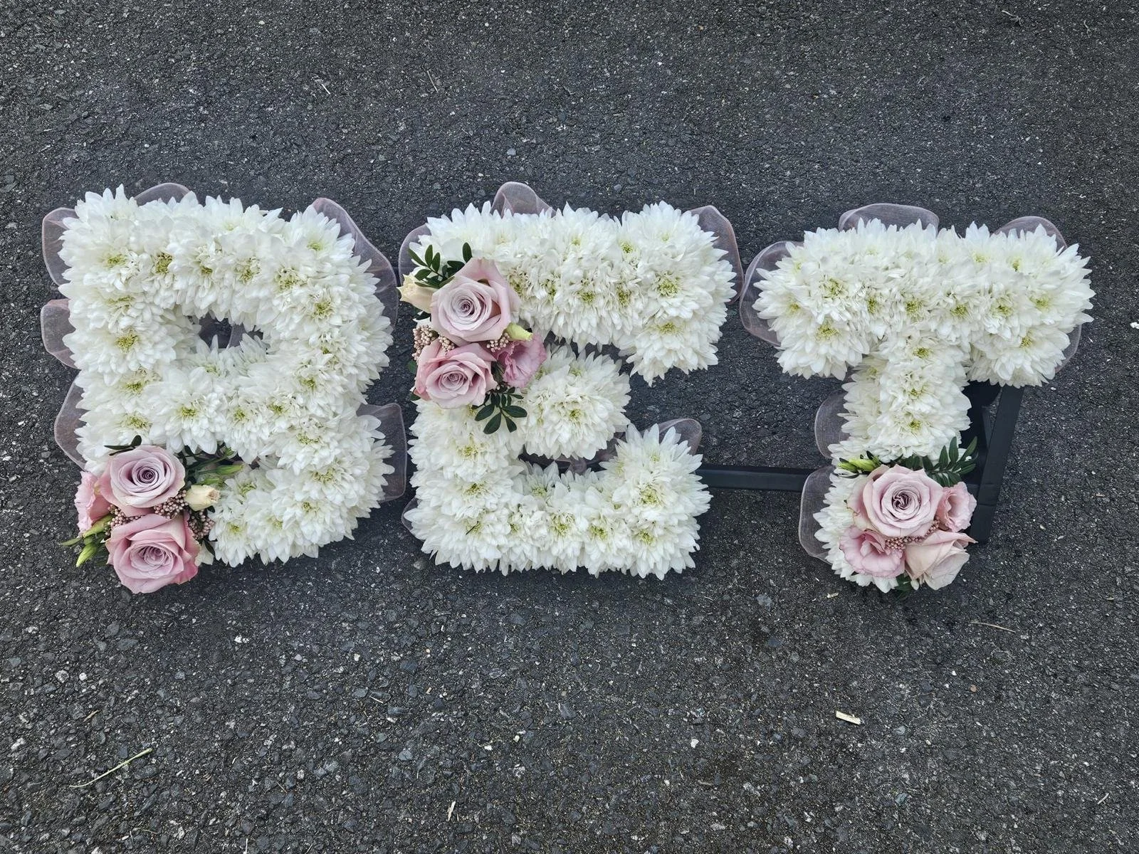 Flowers arranged to spell out the word 'LOVE' on a paved surface, with white flowers for the letters and pink roses with greenery as accents.