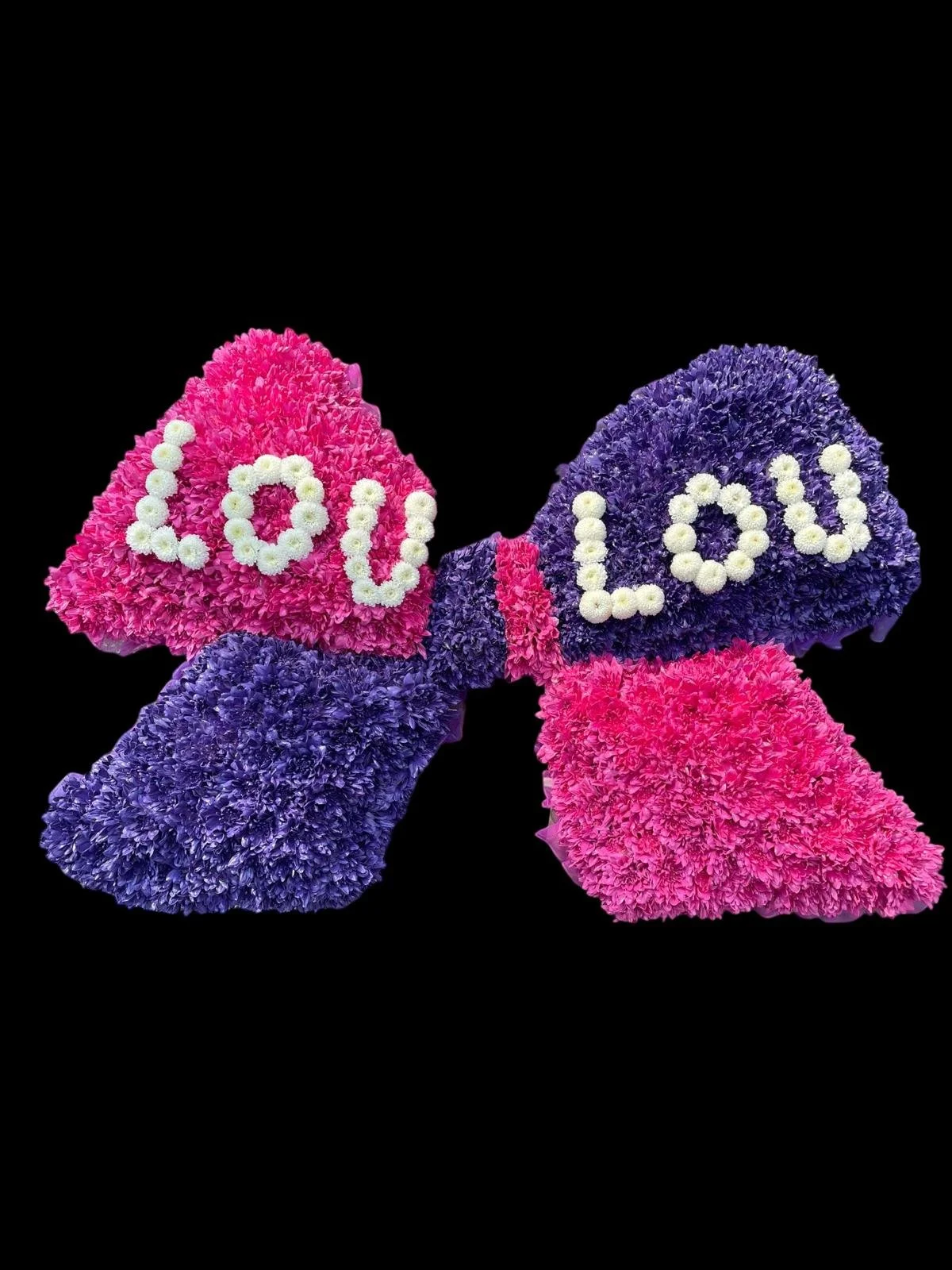 Square arrangement of pink and purple flowers forming a bow with the word 'LOVE' spelled out in white flowers.