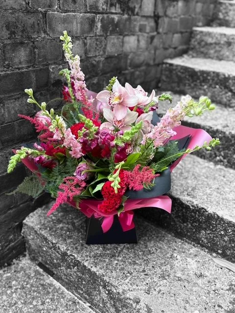A colorful bouquet of pink and red flowers with green foliage, wrapped in pink and black paper, placed on outdoor concrete steps against a brick wall background.