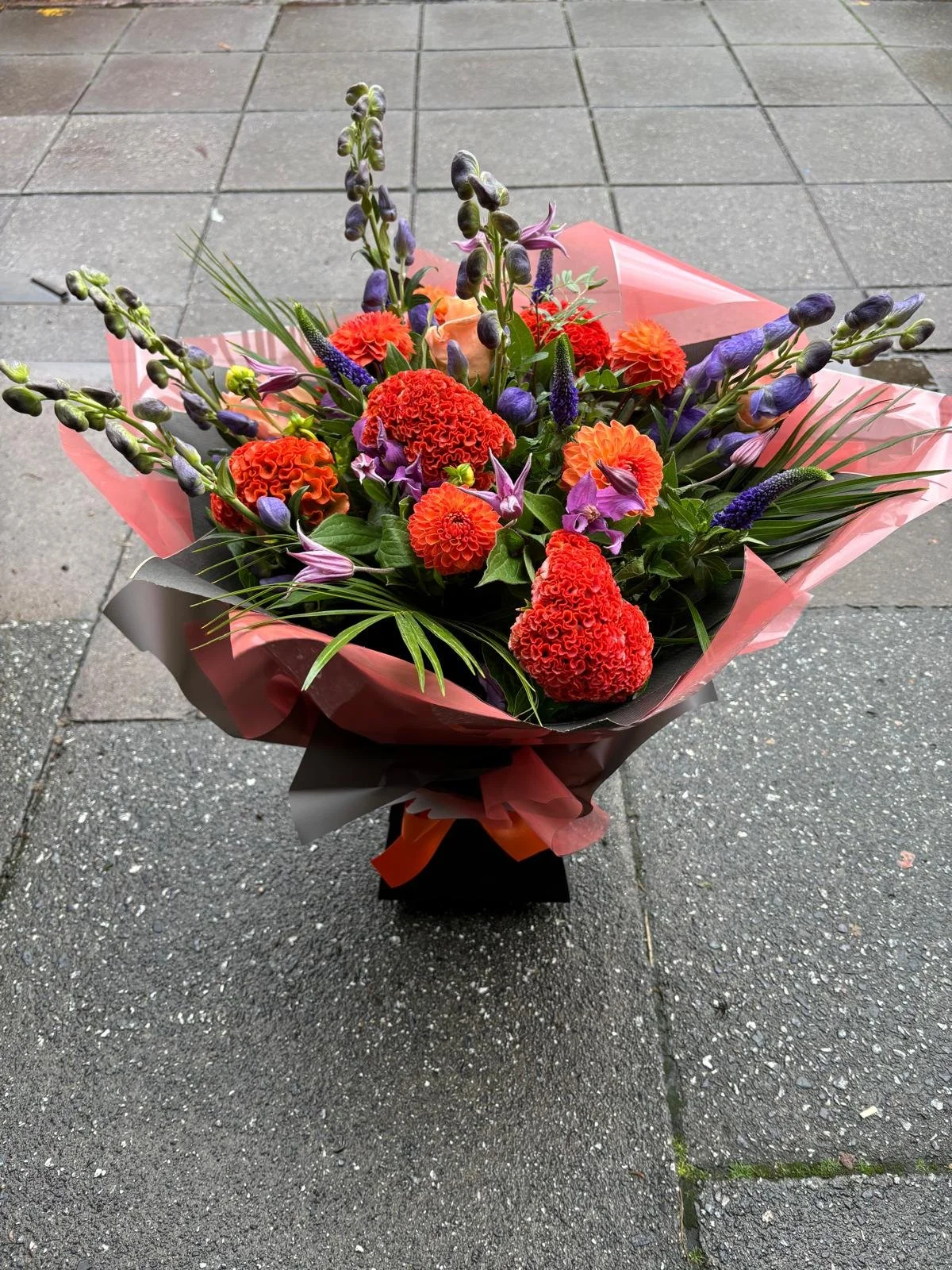 A colorful flower bouquet with red, orange, purple, and green flowers is wrapped in pink and gray paper, placed on a gray sidewalk.