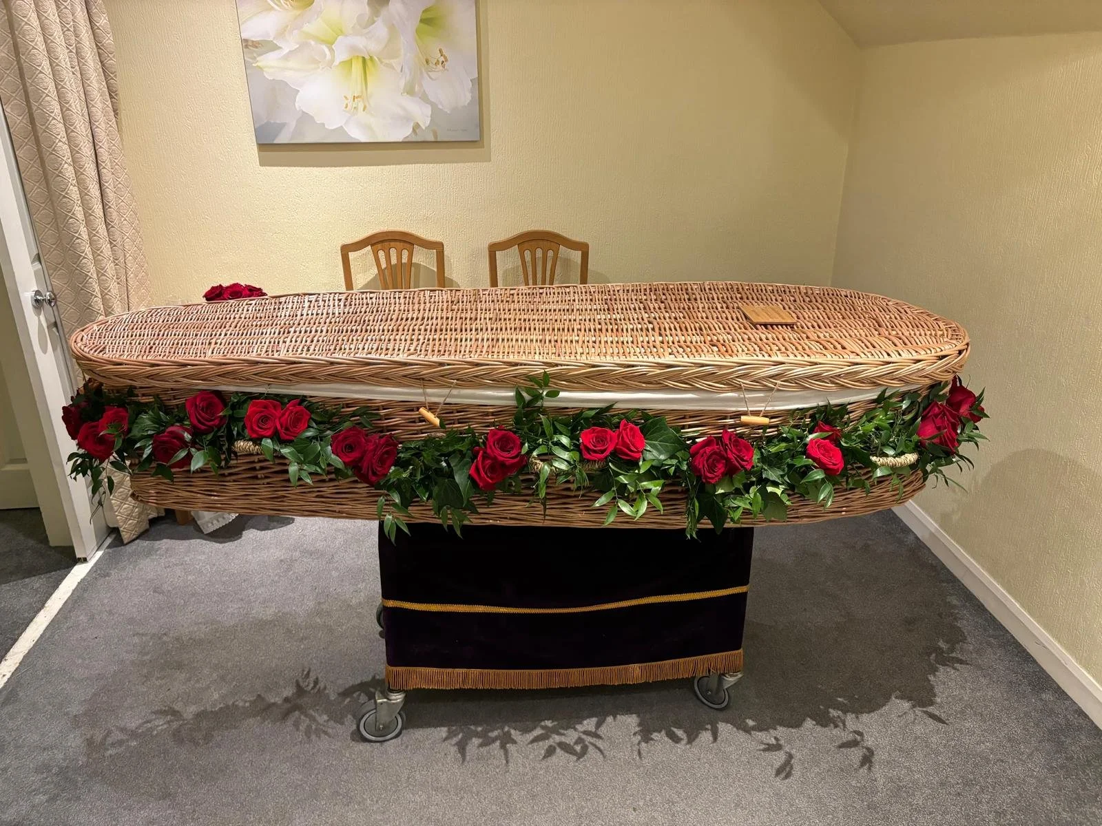 A funeral casket with red roses and green foliage on it, placed on a rolling cart inside a room with beige walls and a small wooden table with two chairs in the background.