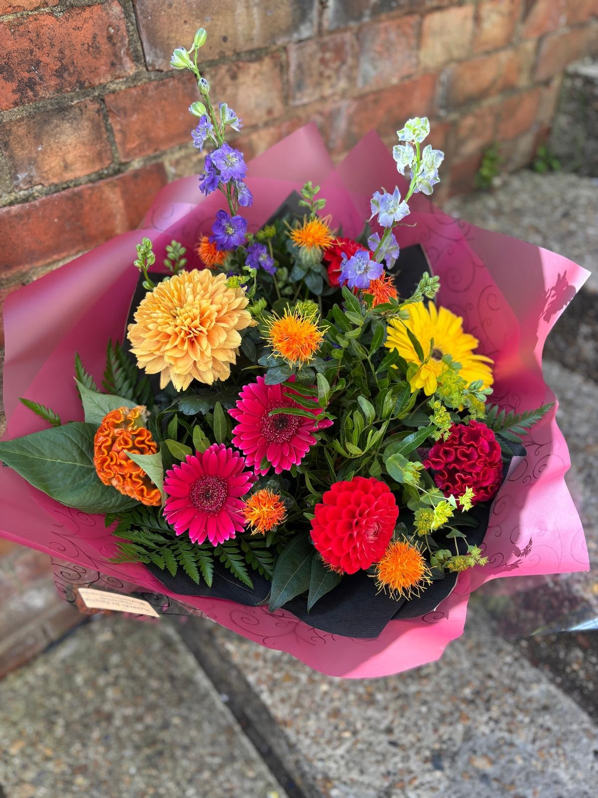 Colorful bouquet of flowers including dahlias, zinnias, marigolds, and other mixed blossoms wrapped in pink paper against a brick wall background.