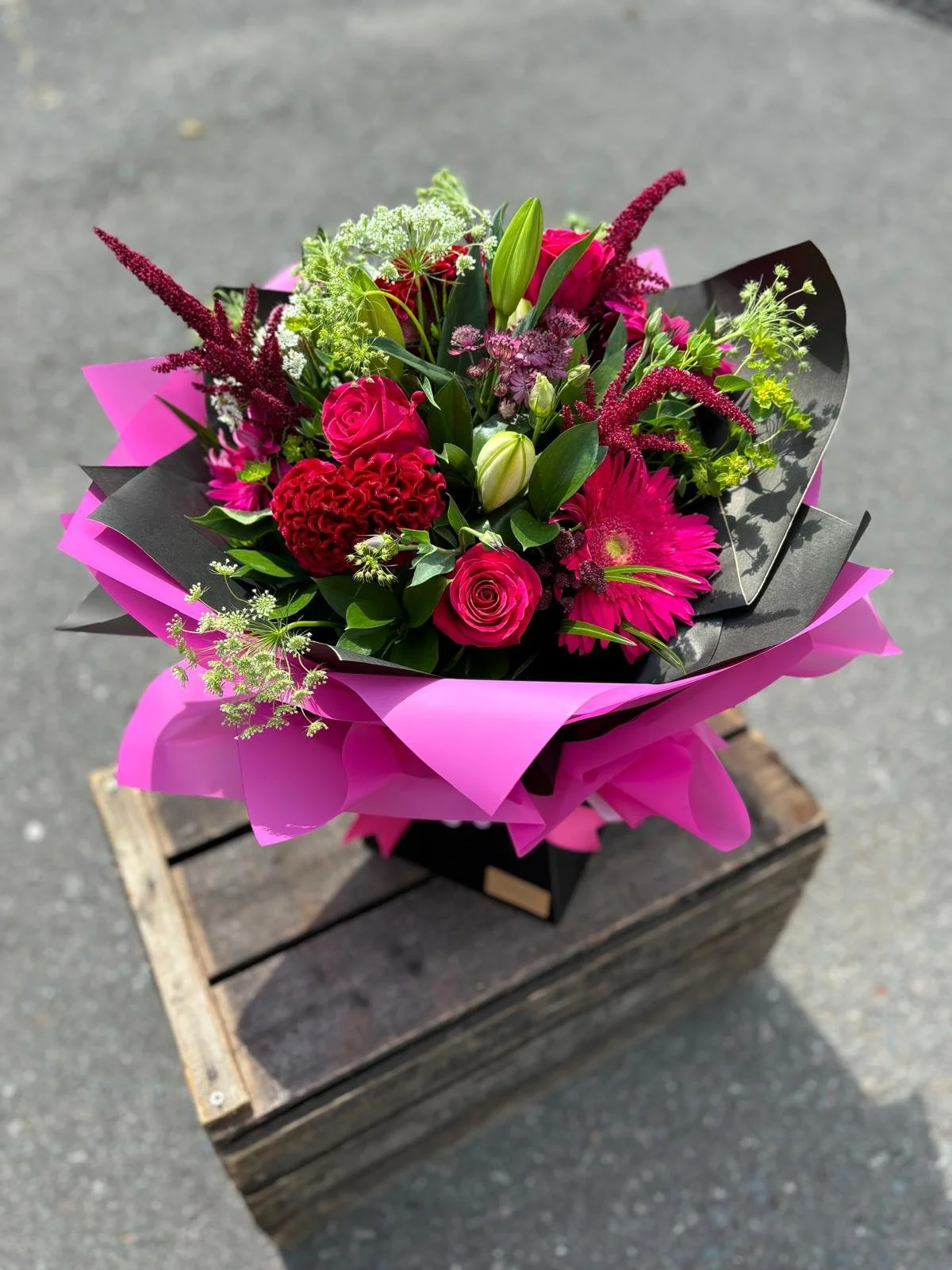 Bouquet of pink, red, and white flowers wrapped in black and pink paper, placed on a wooden stand on a concrete surface.