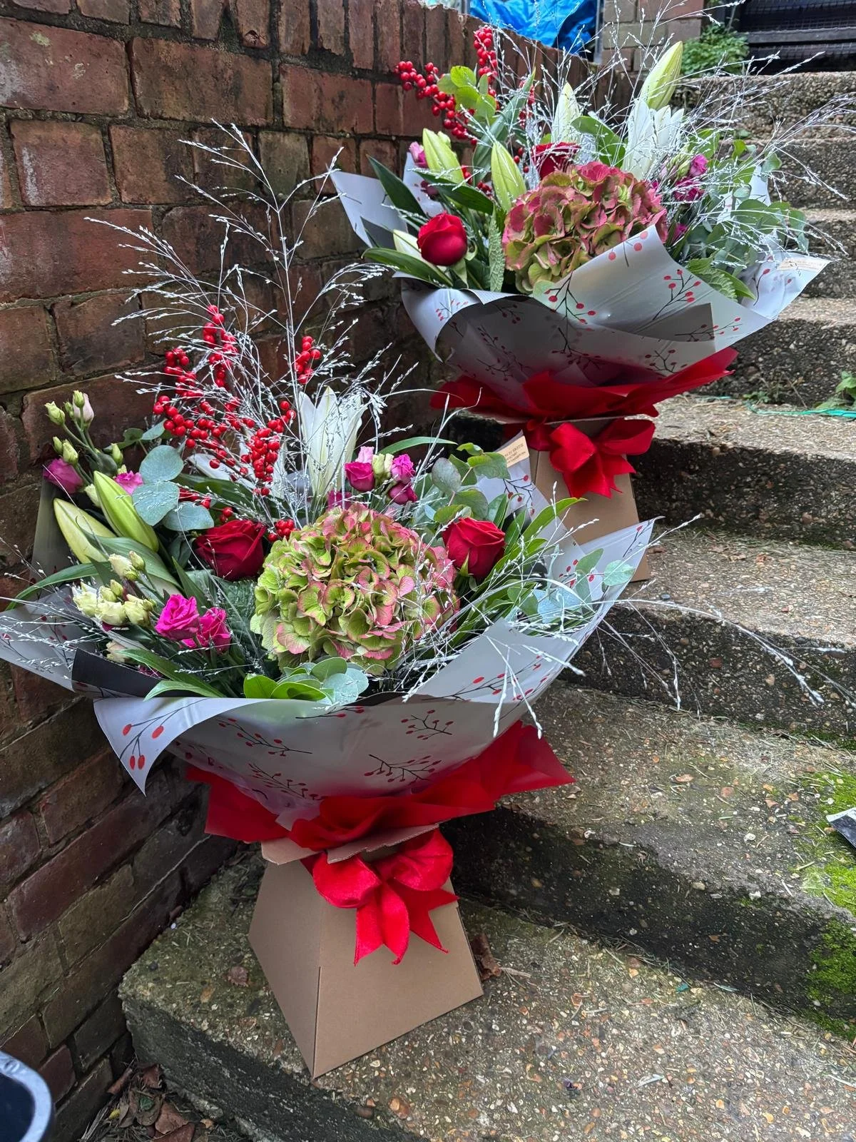 Two floral bouquets placed on outdoor concrete steps against a brick wall. The bouquets contain red roses, pink lisianthus, green hydrangeas, and decorative silver branches, wrapped in white paper with red accents and tied with red ribbons.