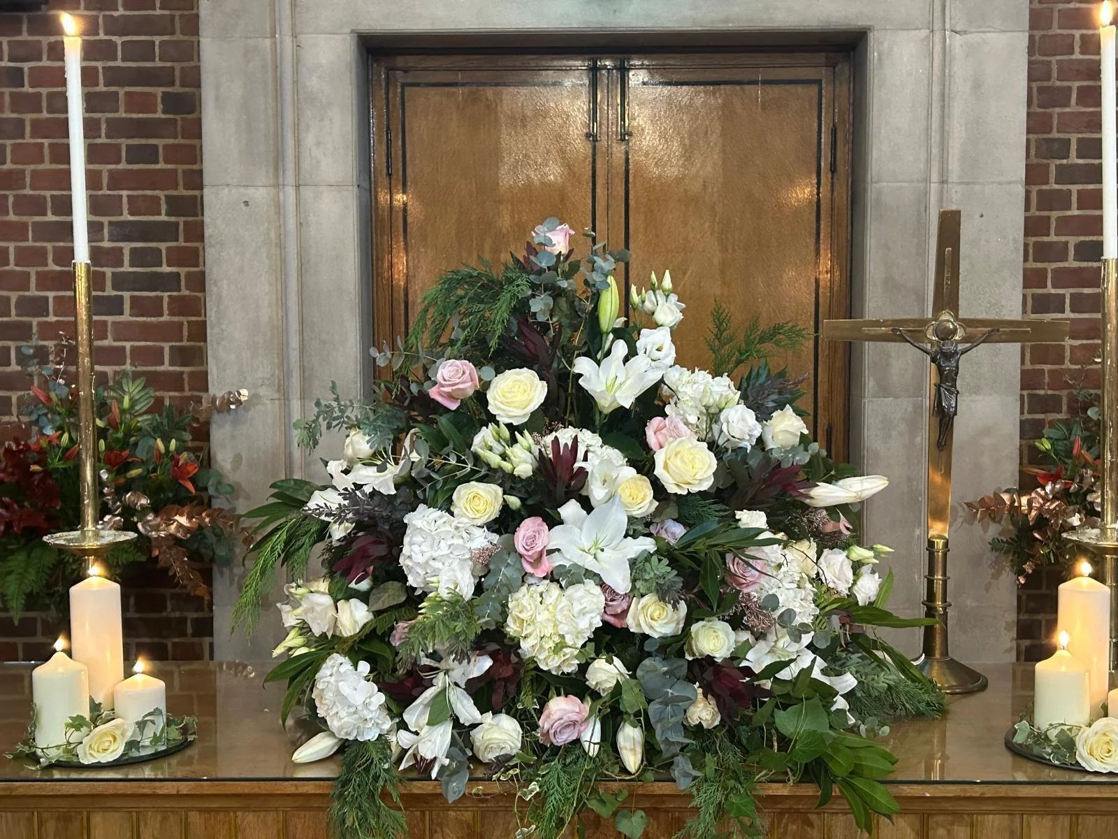 A large floral arrangement with white, pink, and purple flowers, green foliage, candles on either side, and a crucifix on the right, set against the backdrop of a church altar with a gold door and brick walls.