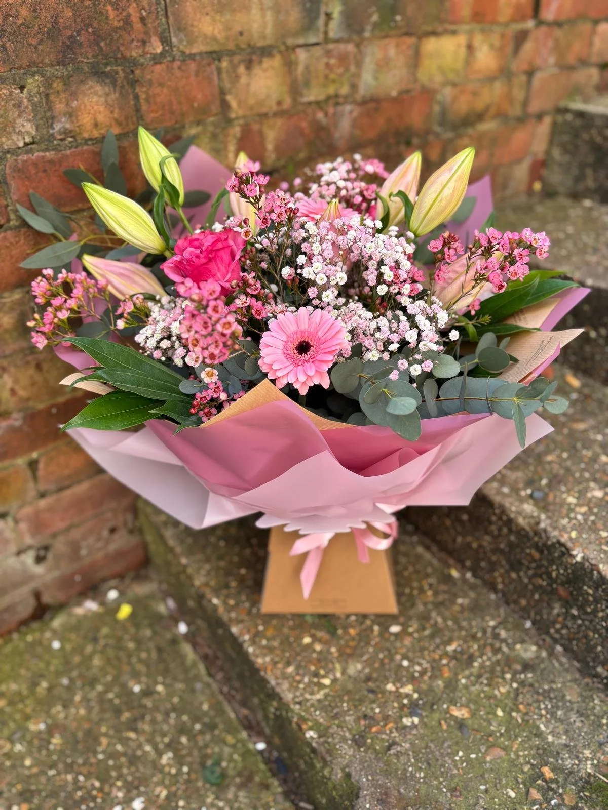 A bouquet of pink and white flowers including roses, gerbera daisies, and baby's breath, wrapped in pink paper, with green eucalyptus leaves, placed outdoors against a brick wall.