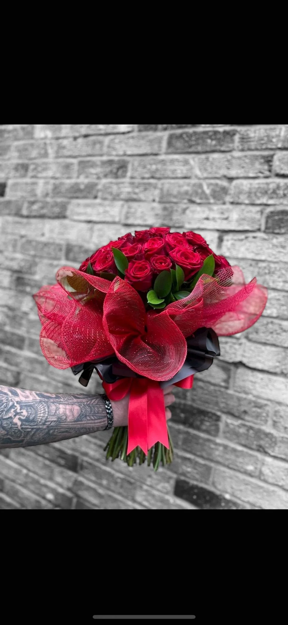 Hand holding a bouquet of red roses wrapped with red and black mesh and ribbons, against a gray brick wall.