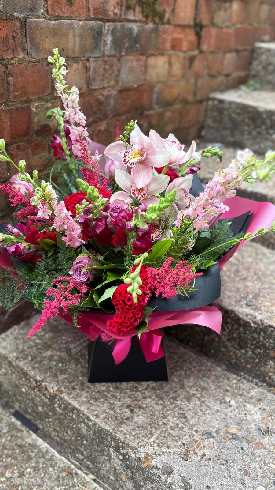 A vibrant bouquet of pink, red, and purple flowers wrapped in pink and black paper, with a pink ribbon, placed on concrete steps against a brick wall background.