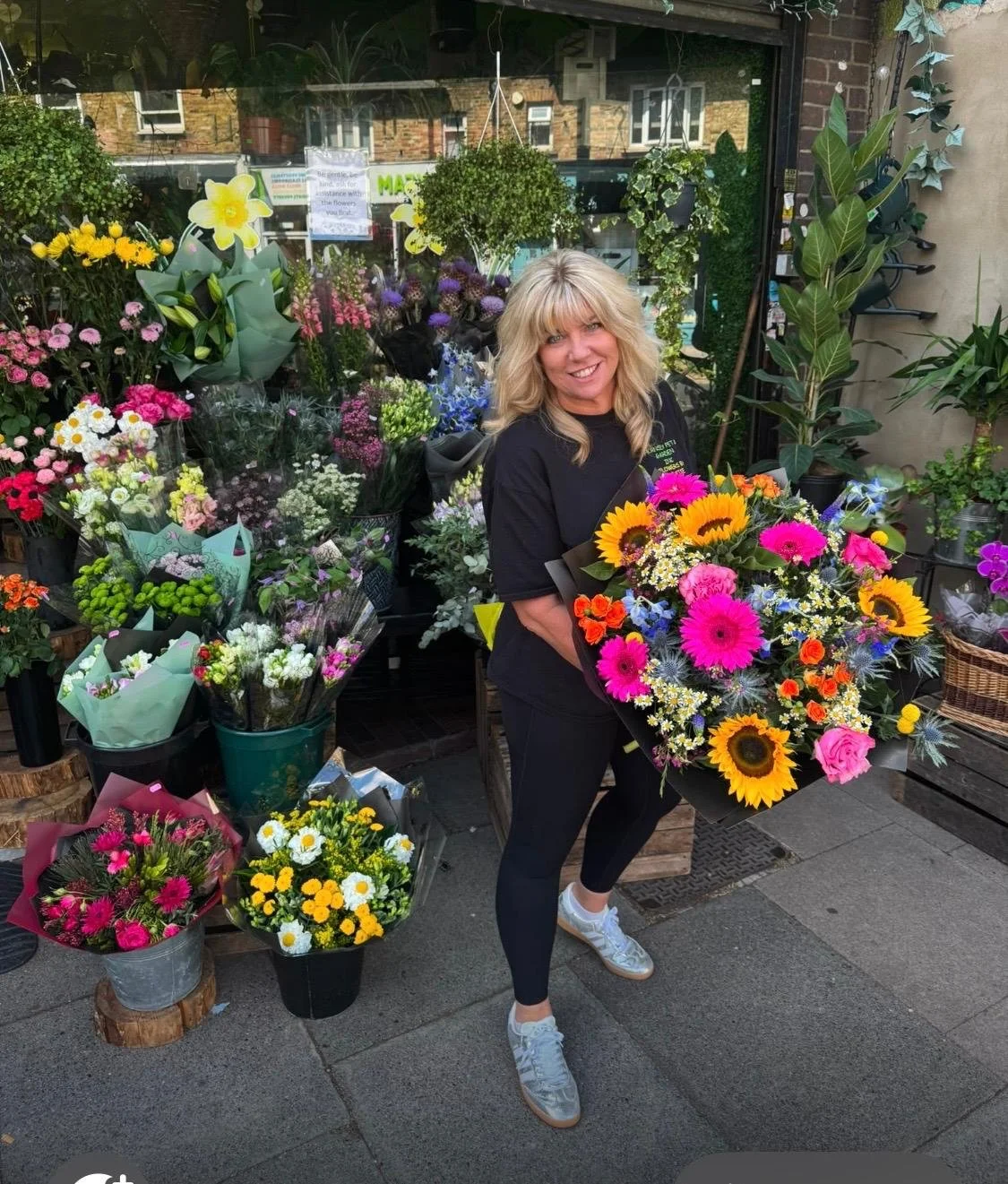 A woman holding a large colorful bouquet of flowers stands outside a flower shop, surrounded by various other flower arrangements and bouquets.