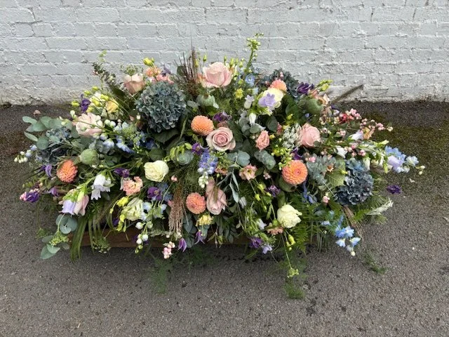 A long, colorful floral arrangement with pink roses, blue hydrangeas, orange and white blossoms, and green foliage, placed on the ground against a white brick wall.