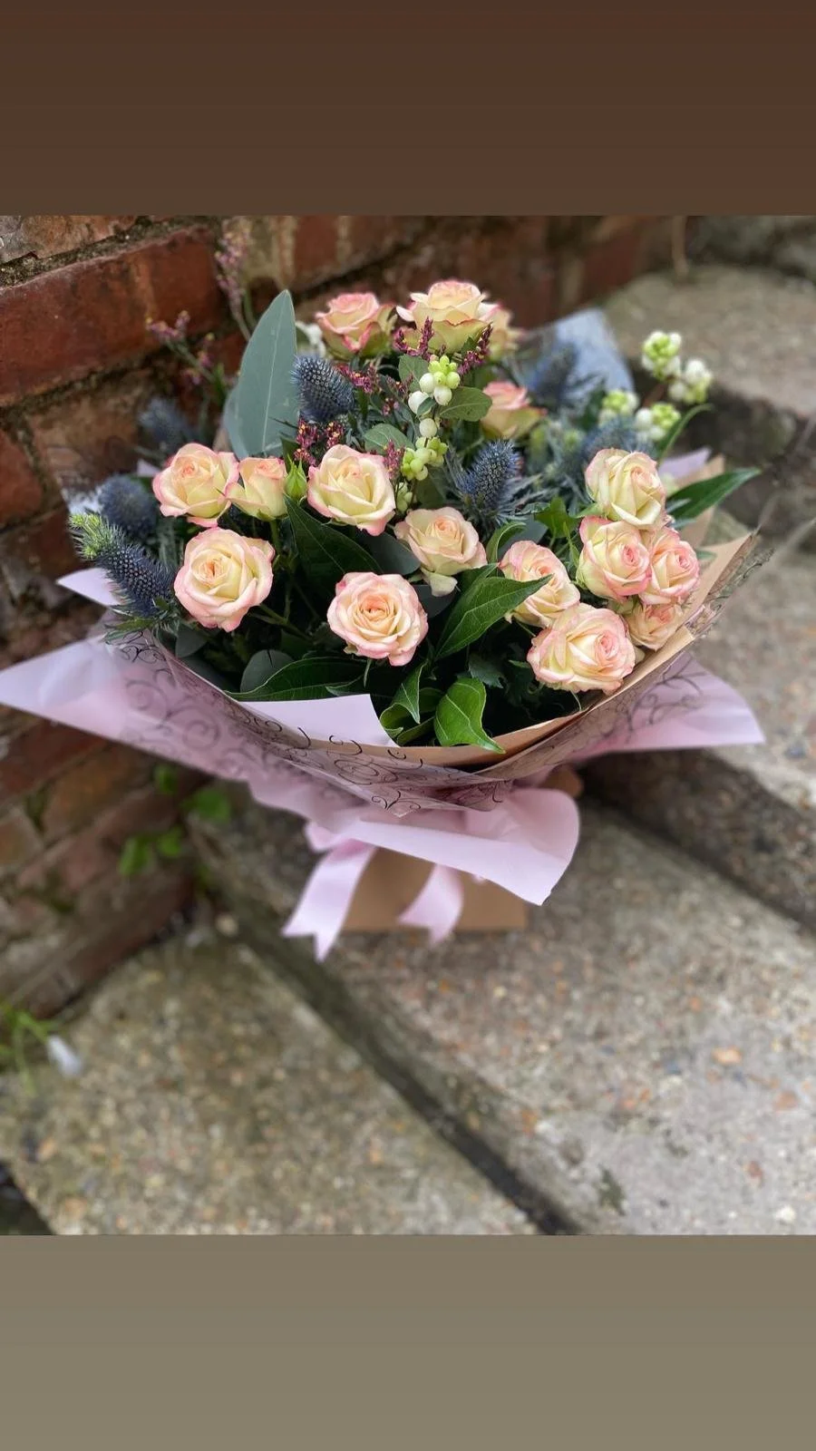 A bouquet of light pink roses, green foliage, and blue thistle flowers wrapped in pink and beige paper, resting on a brick ledge.