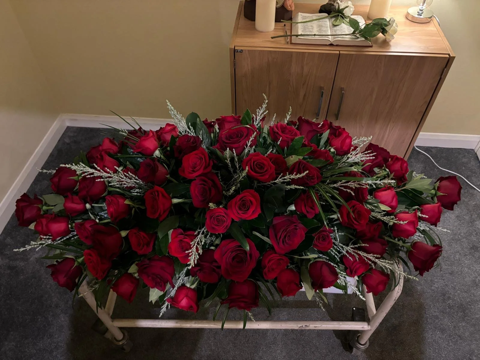A table covered with a large bouquet of red roses with green leaves and white filler flowers, placed in a white container.