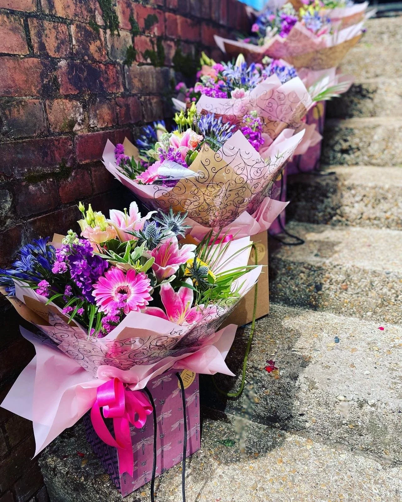 Multiple colorful flower bouquets wrapped in pink and decorative paper are lined up against a red brick wall on the ground.