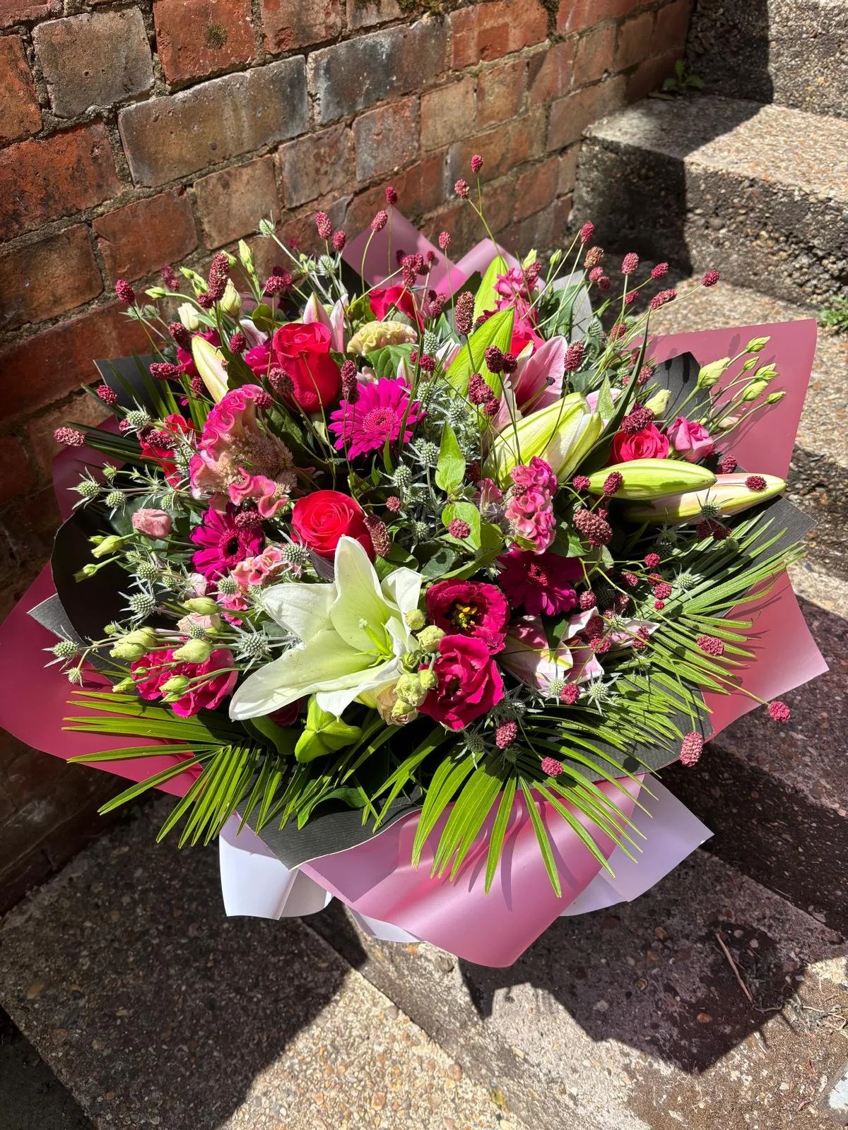 A vibrant bouquet of pink, white, and green flowers including lilies and roses, wrapped in pink and black paper, resting against a brick wall on a concrete sidewalk.
