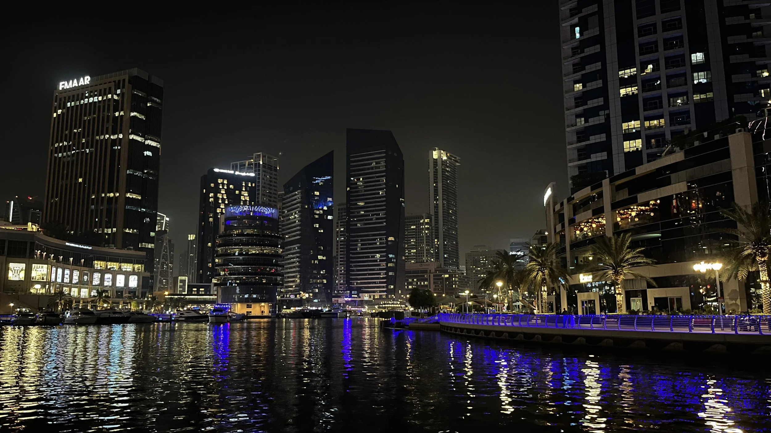 Blick auf eine beleuchtete Stadt bei Nacht mit Wasser im Vordergrund, Hochhäuser und Palmen