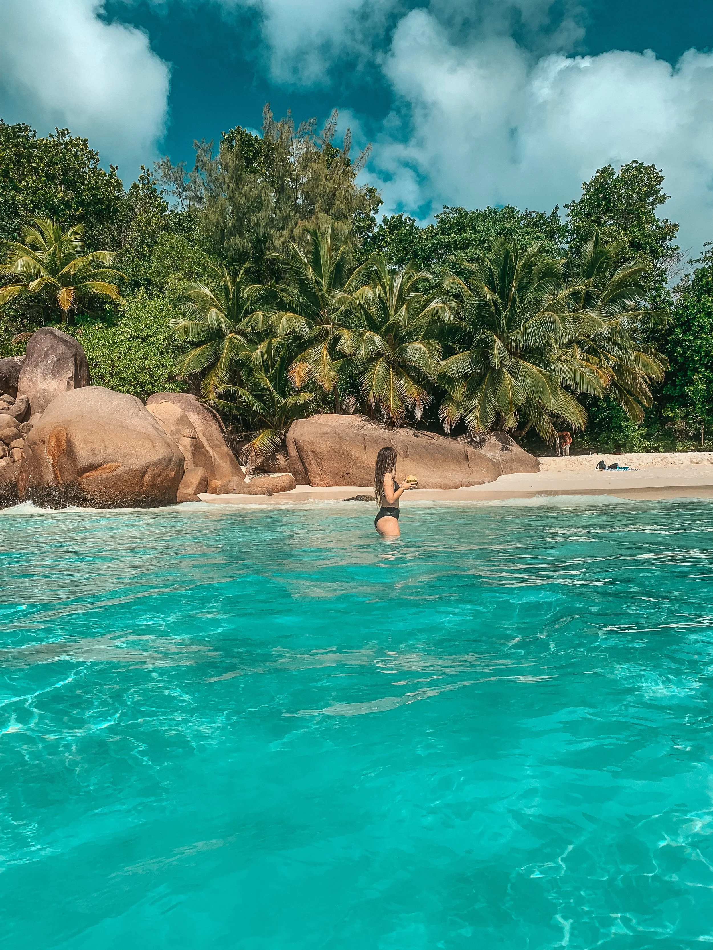 Frau steht im Wasser am Strand, umgeben von großen Felsen, Palmen und grünen Bäumen, mit einem blauen Himmel und Wolken im Hintergrund.