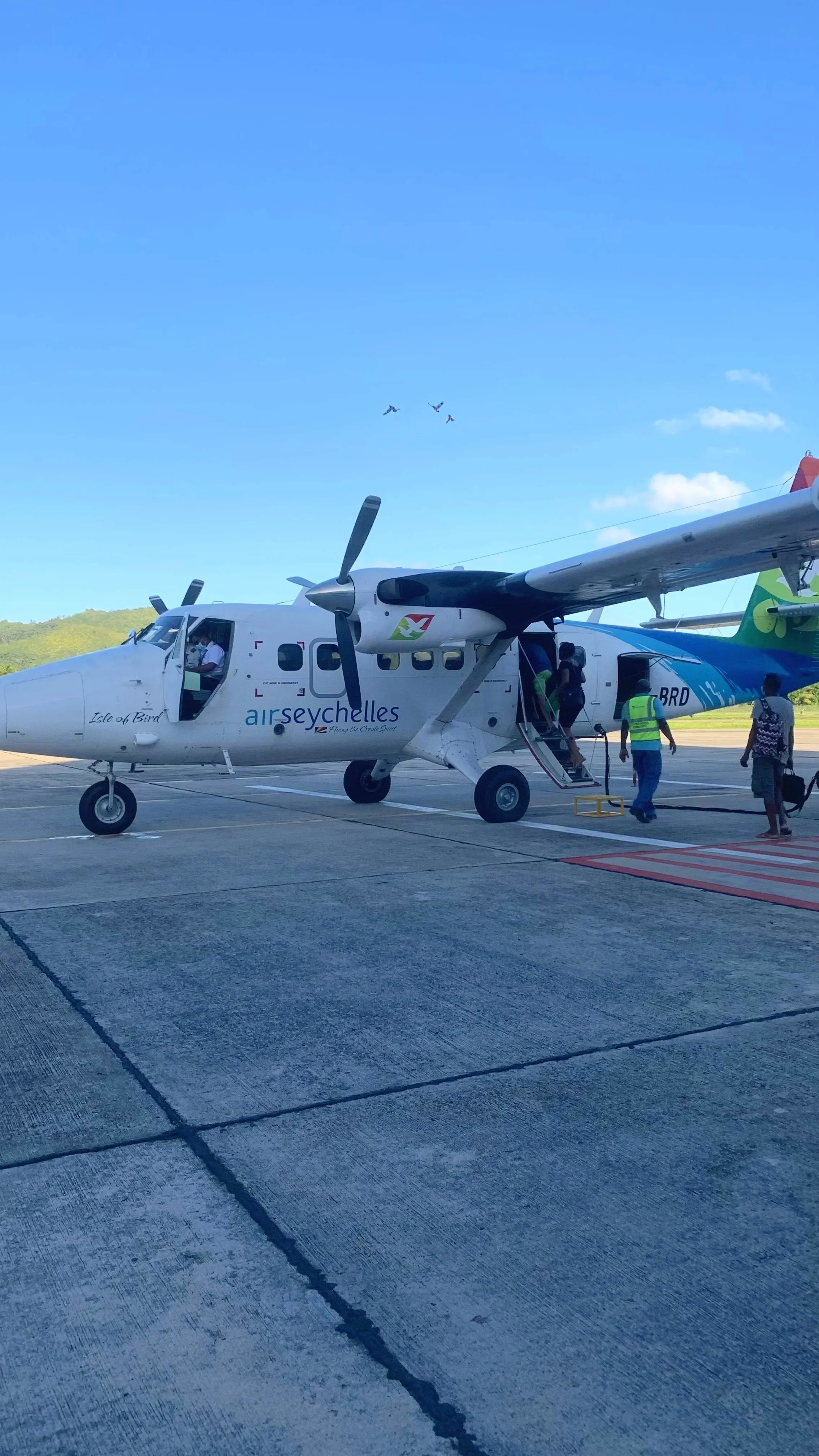 Ein kleines Passagierflugzeug auf einem Rollfeld, das für den Flug vorbereitet wird, mit Flugbegleitern und Passagieren beim Einsteigen, während Vögel am Himmel fliegen.